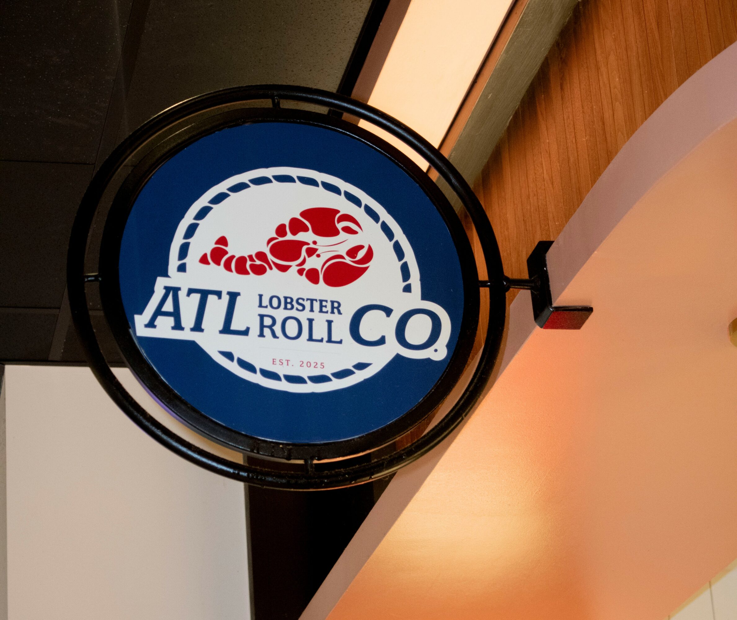 Round sign with a lobster illustration and text 'ATL Lobster Roll Co' hanging from a ceiling, with warm lighting.