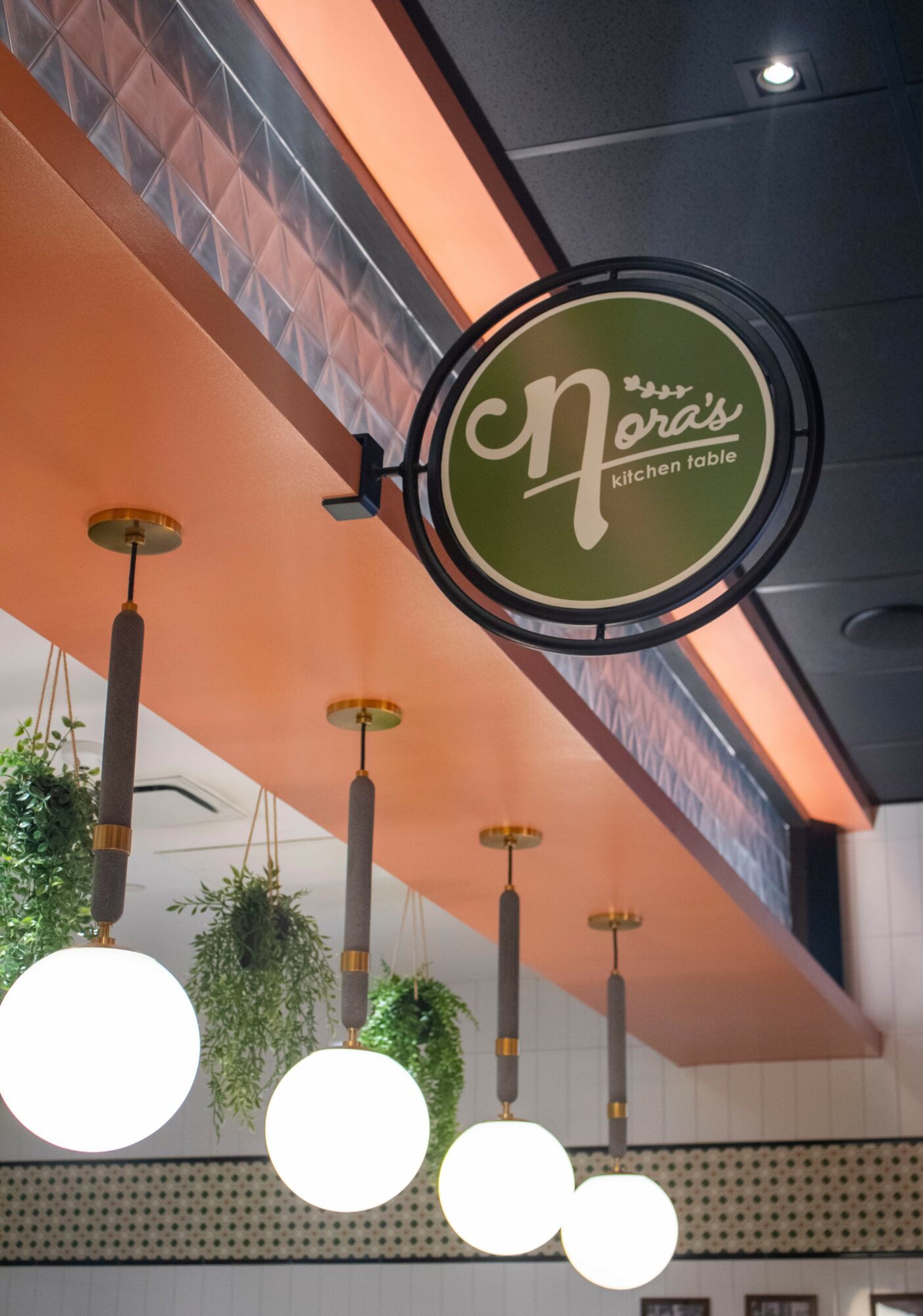 Restaurant sign hanging from ceiling with four hanging lights and plants below, interior with orange and black ceiling accents.