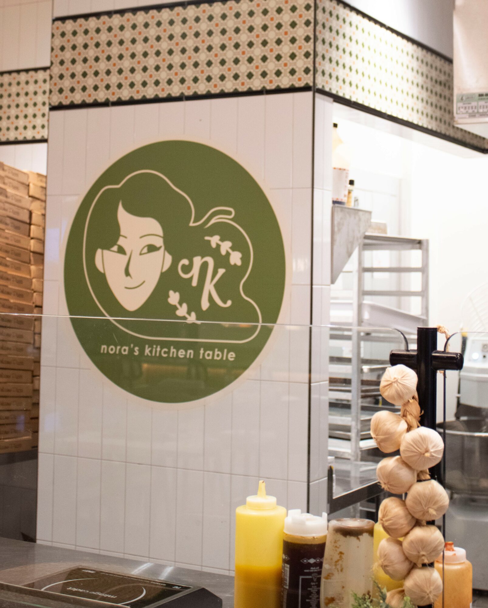 Wall with logo of a woman's face and text 'nora's kitchen table', kitchen equipment visible in background.