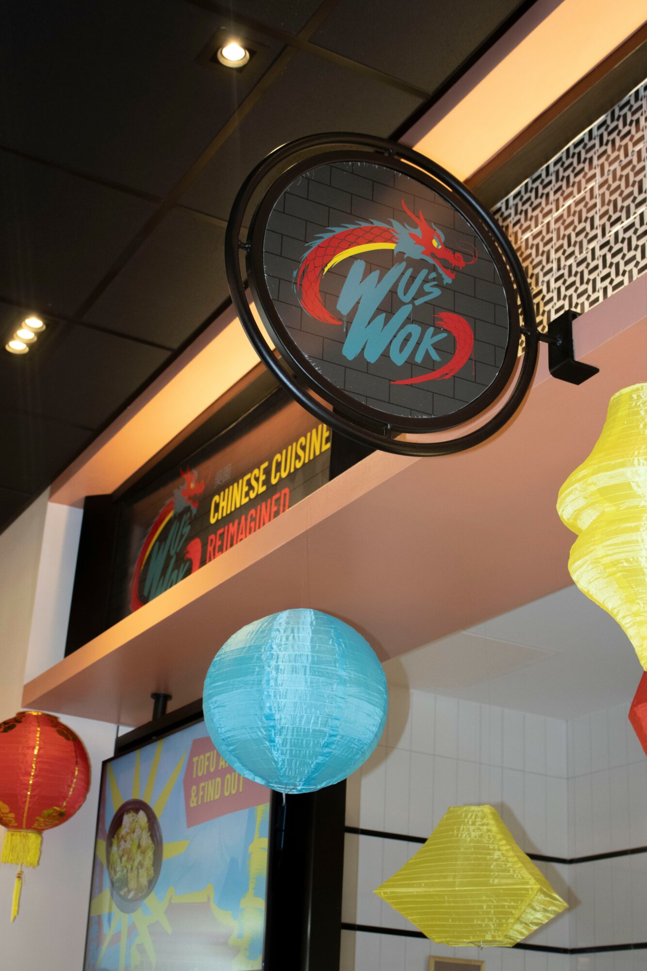 Circular sign with colorful text and graphics, hanging from ceiling, with paper lanterns below, in an indoor setting.
