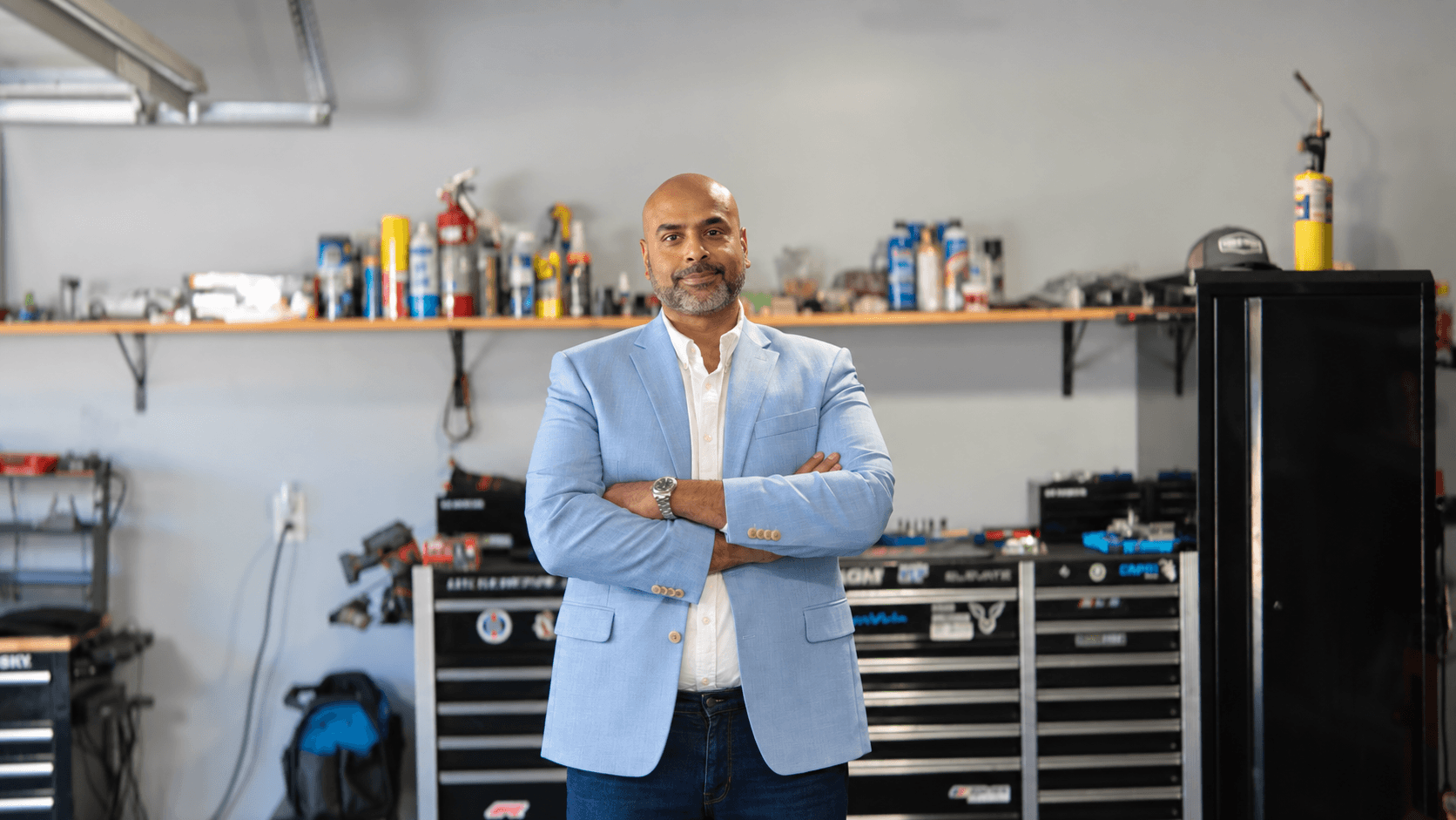 Man in a light blue blazer standing with arms crossed in a workshop or garage, with tools and supplies on shelves behind him.