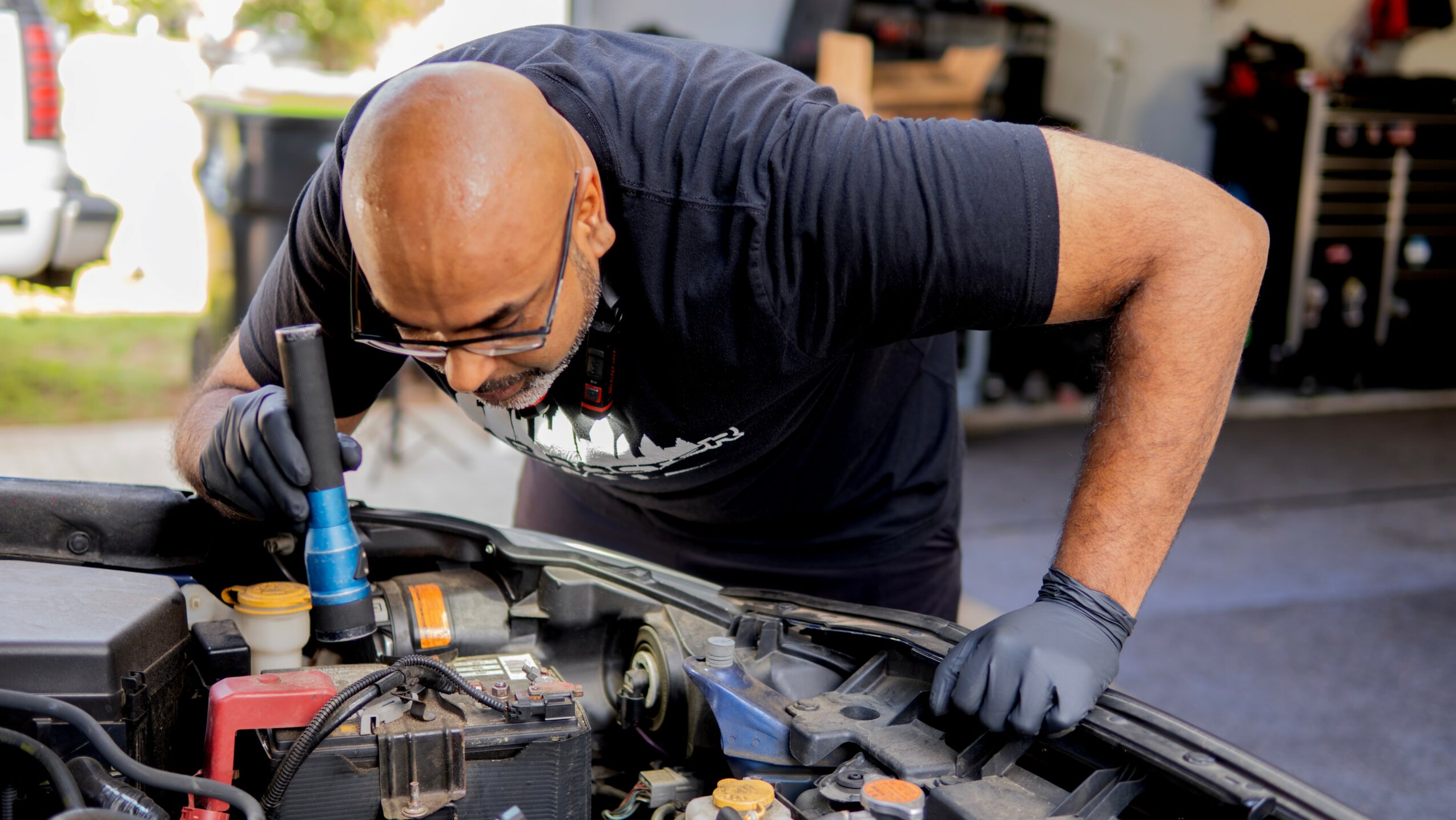 Man wearing glasses and gloves working on car engine in garage, leaning over engine bay.