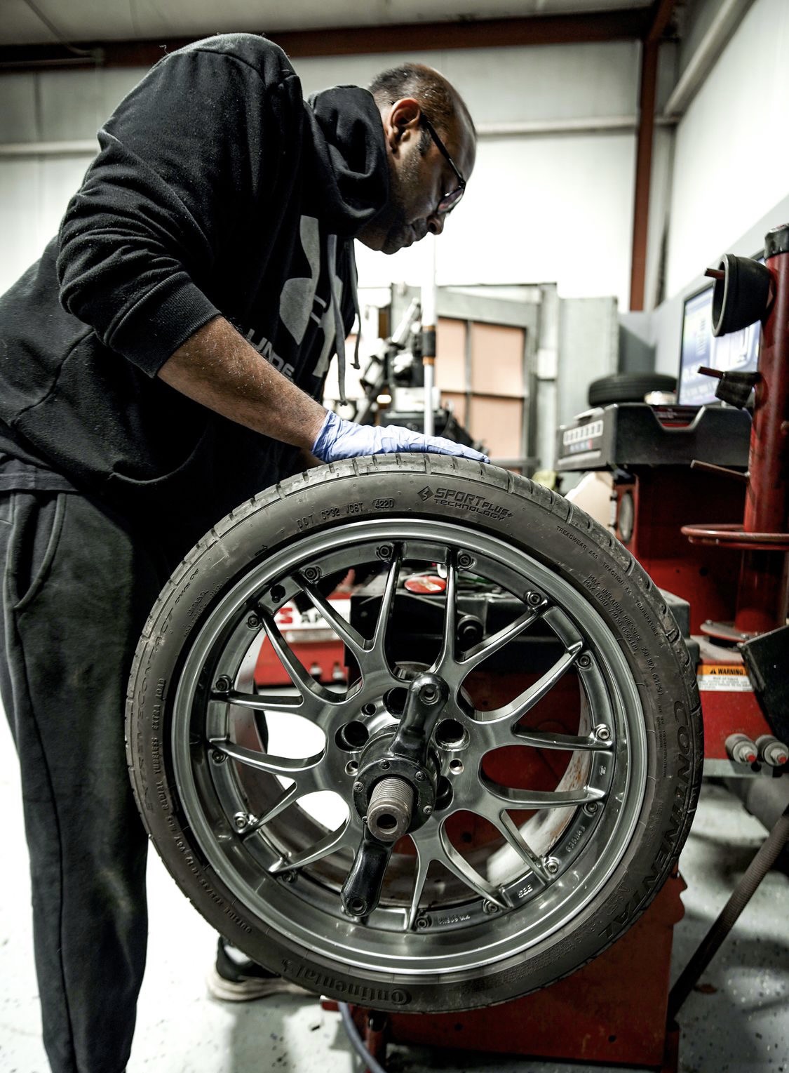 Person working on a car wheel in a garage, wearing glasses and gloves, with tools and equipment around.