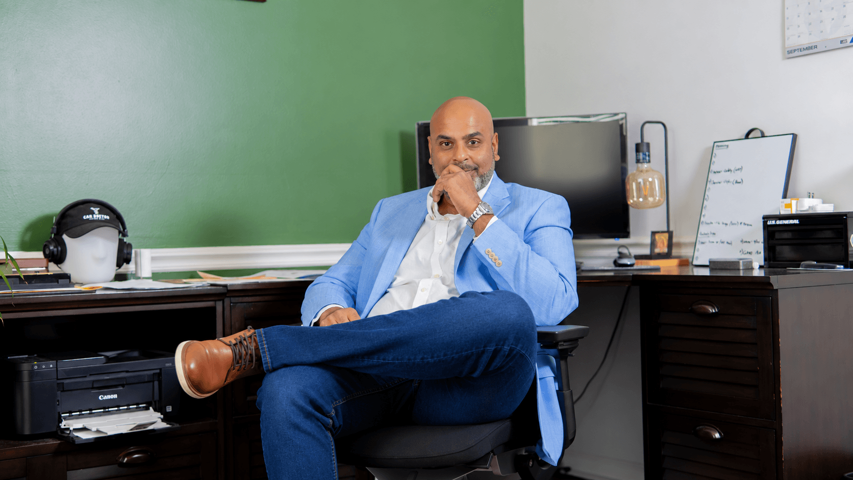 Man in blue suit sitting at desk with hand on chin, office setting with computer and equipment behind him.