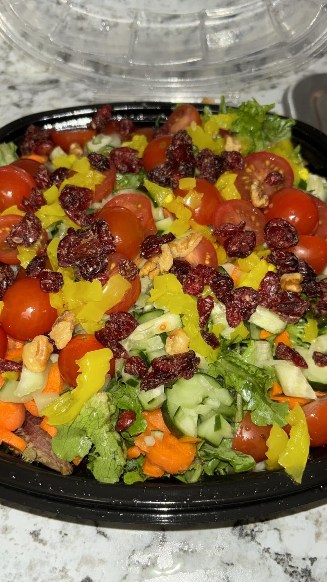 Mixed salad with cherry tomatoes, cucumber, yellow bell peppers, lettuce, and dried cranberries in a black container.