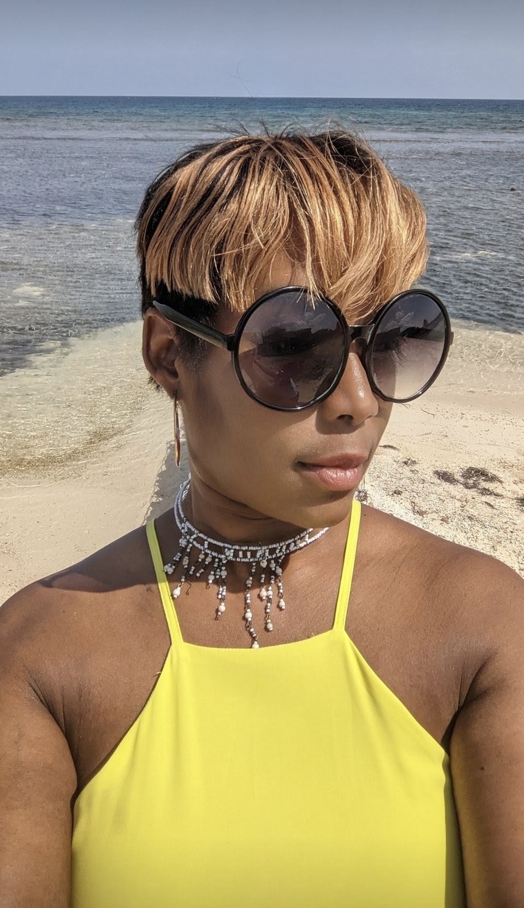 Woman with short hair wearing large sunglasses and a yellow top at the beach.