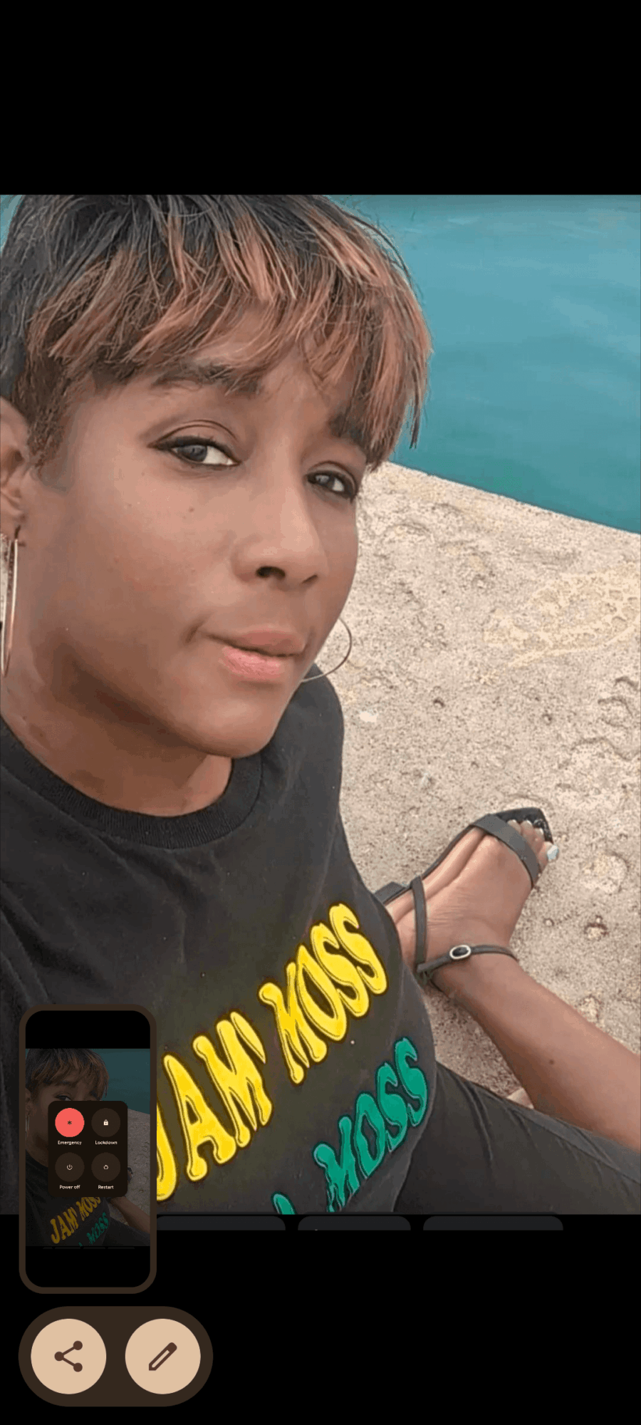 Person with short hair sitting on a beach, wearing a black shirt with colorful text, near water and sand.