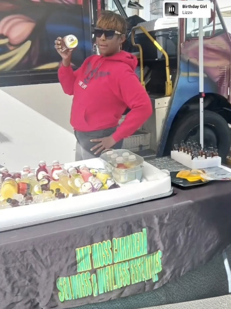 Child in red hoodie holding a bottle at a food stall with colorful bottles and a bus in background.