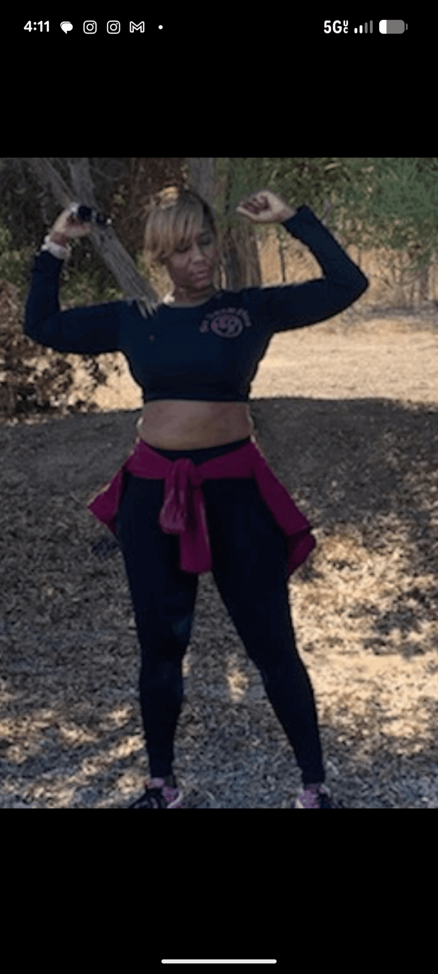 Person standing outdoors with arms raised, wearing a black crop top and leggings, with a maroon jacket tied around waist.