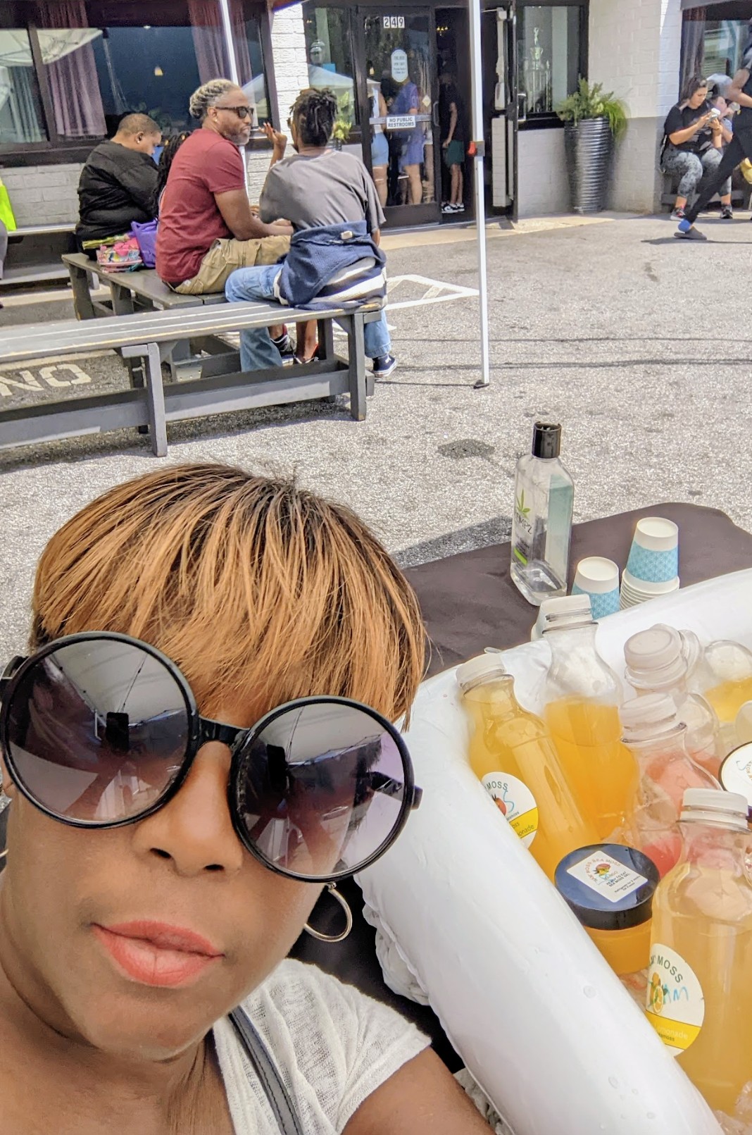 Person with large sunglasses taking selfie outdoors, table with drinks and supplies in foreground, people sitting on benches in background.
