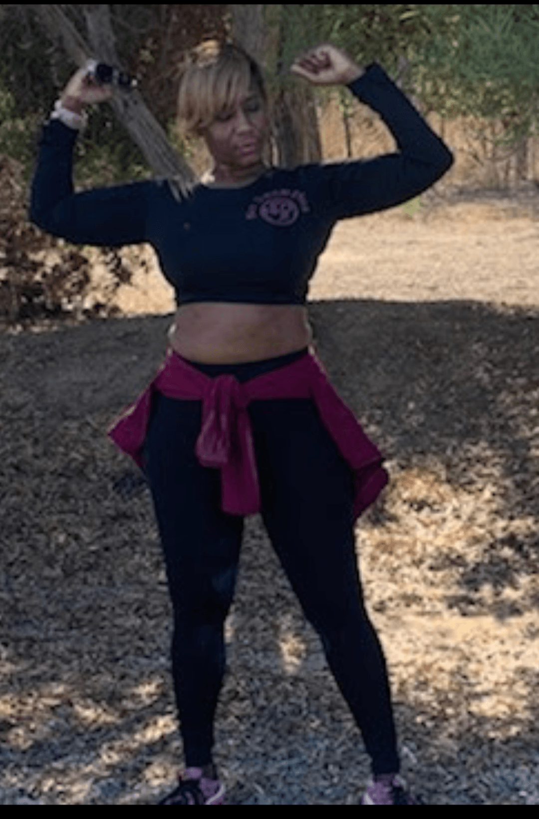 Person standing outdoors with arms raised, holding a baseball bat, wearing a black crop top and dark leggings.