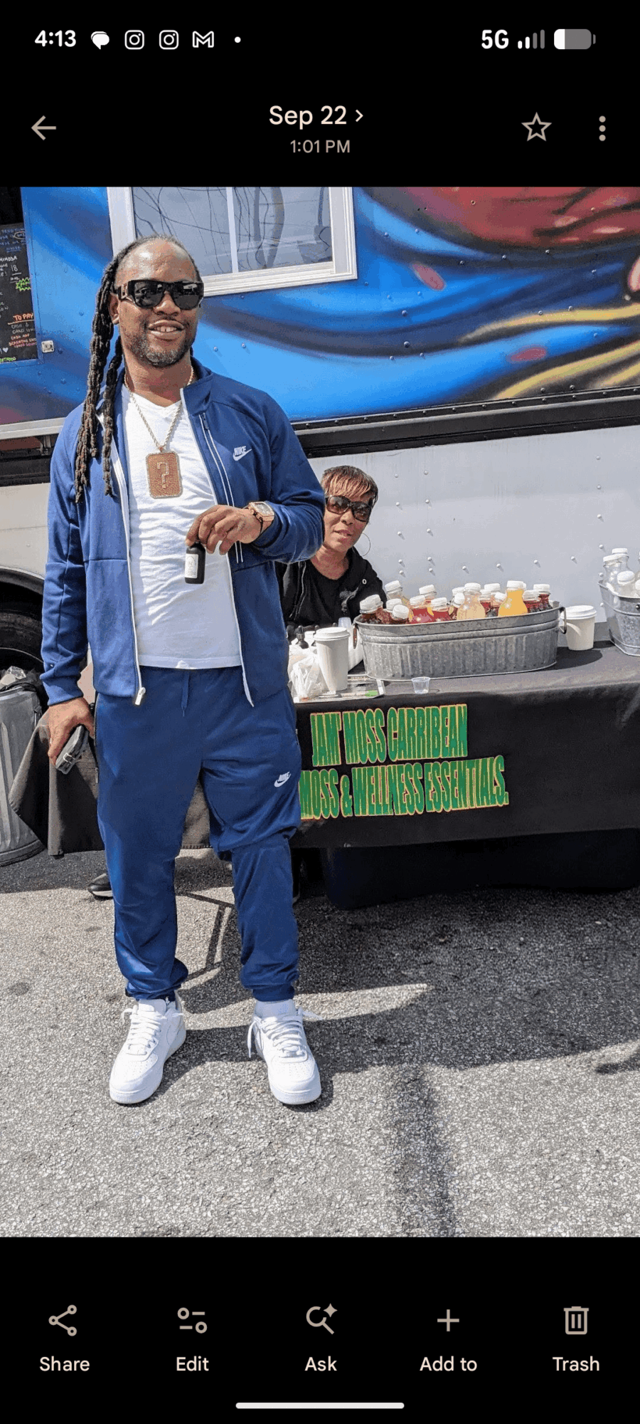 Man with sunglasses and dreadlocks standing outdoors, with a young boy and a table of food in the background.