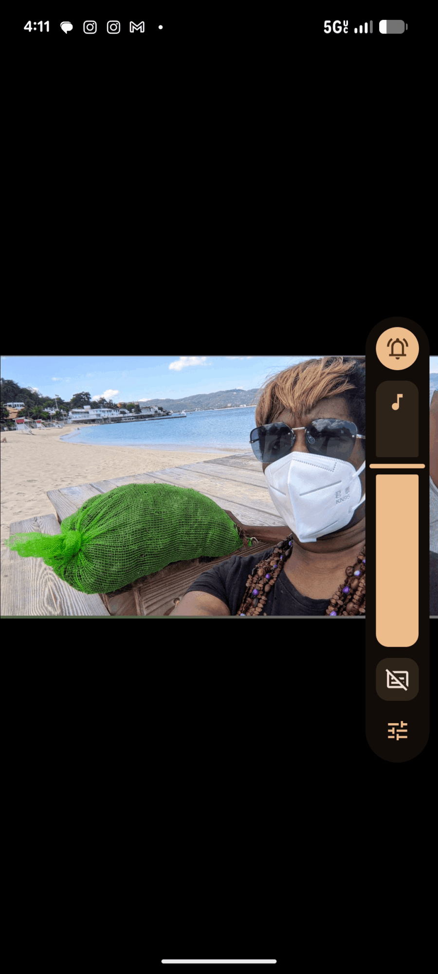 Person wearing sunglasses and a face mask at the beach with a green turtle-shaped pillow, ocean and sky in background.