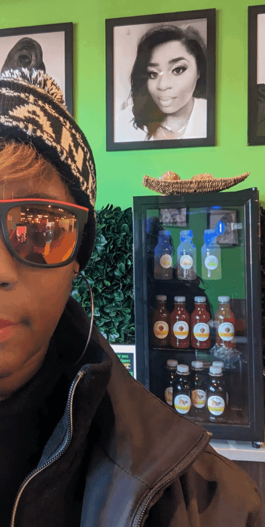 Part of a person's face wearing sunglasses and a beanie, with framed black-and-white portraits on a green wall behind.