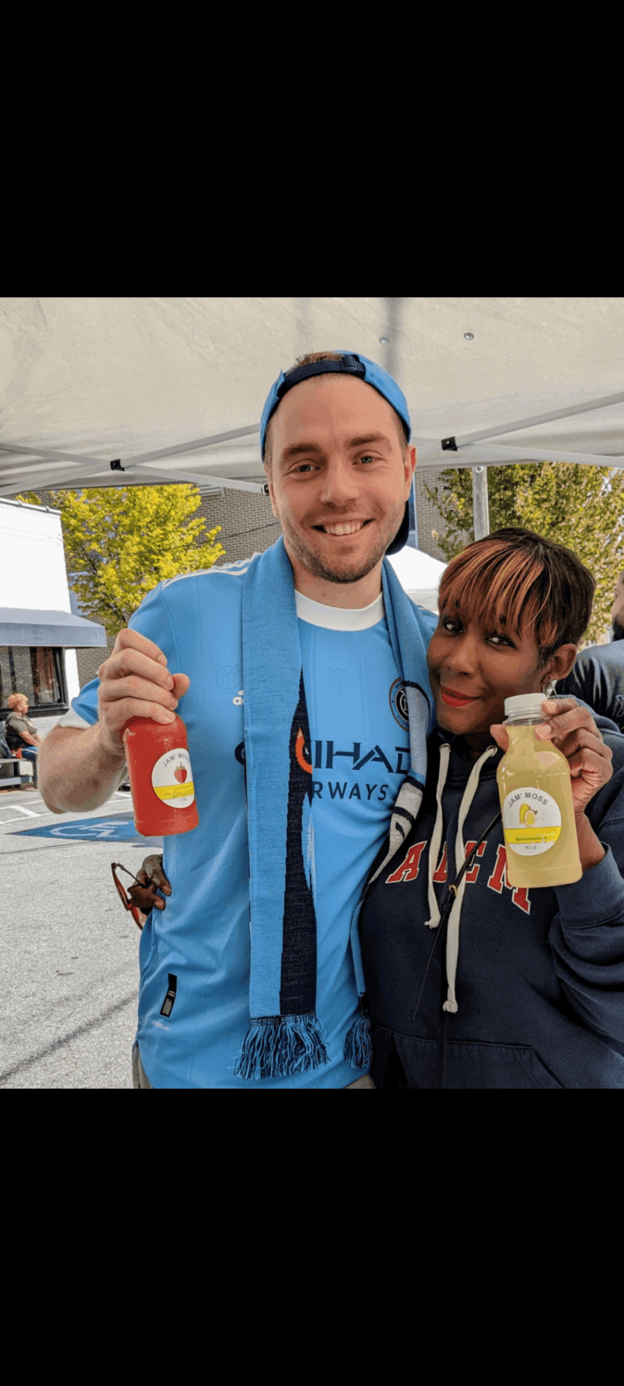 Two people smiling and holding bottles of drinks outdoors, with a tent and trees in the background.