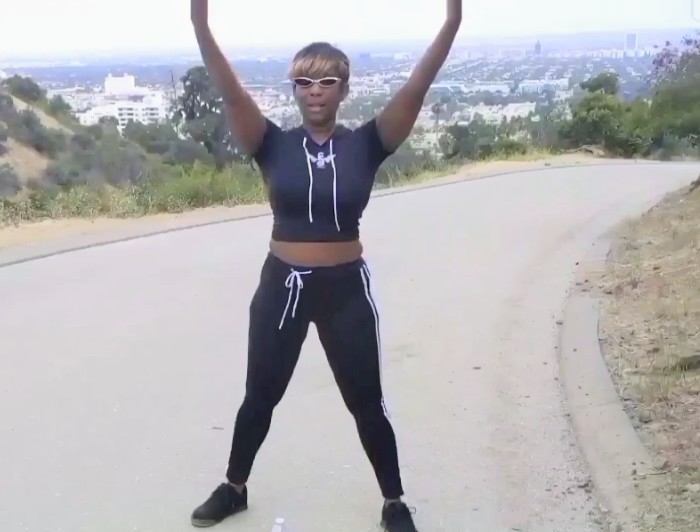 Person standing on a curved road with arms raised, wearing sunglasses, a black athletic outfit, and sneakers, outdoors with cityscape in background.