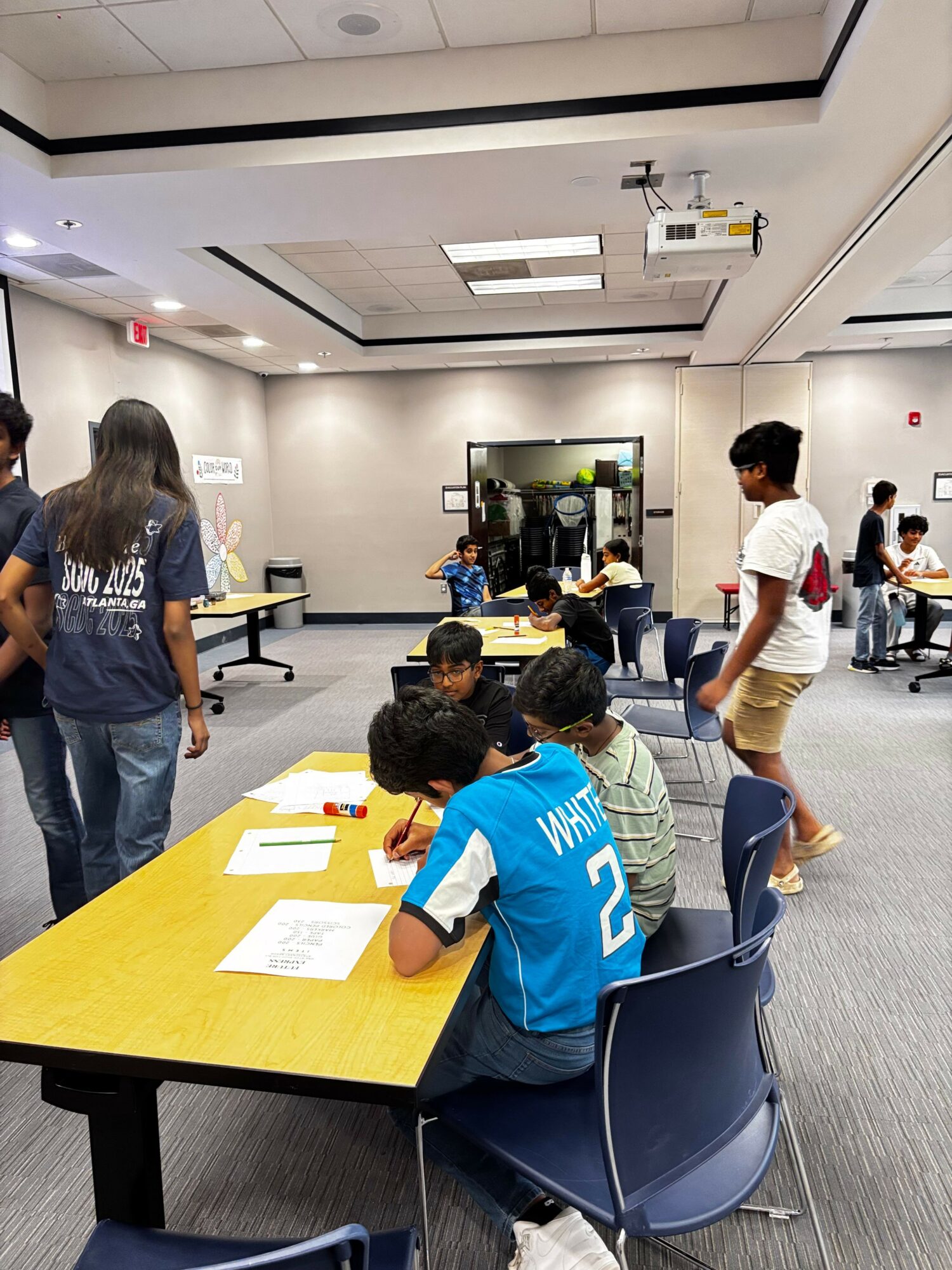 People in a waiting area with chairs, tables, and papers, some standing and some seated, in a modern indoor space.