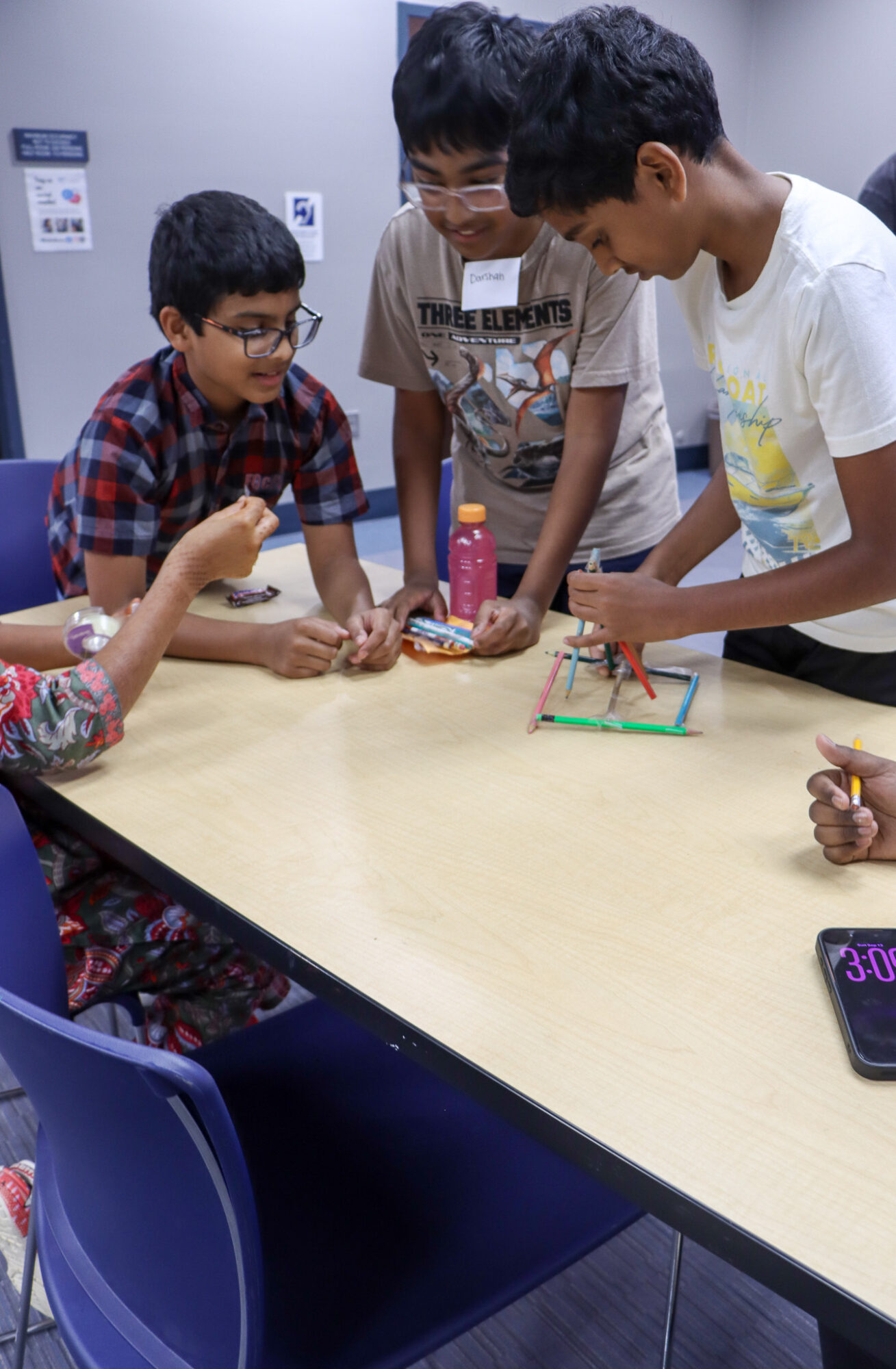 Group of children gathered around a table playing a board game in a room with blue chairs and a gray wall.