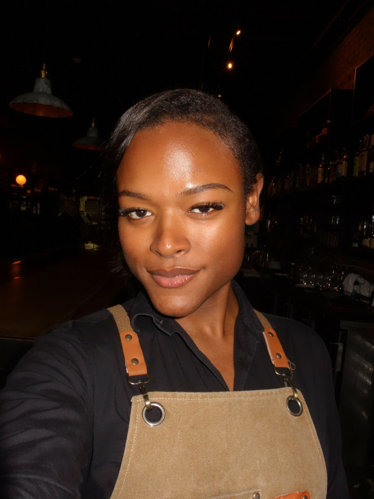 Young woman with dark hair and makeup, wearing a black top and tan apron, smiling in a nighttime setting.