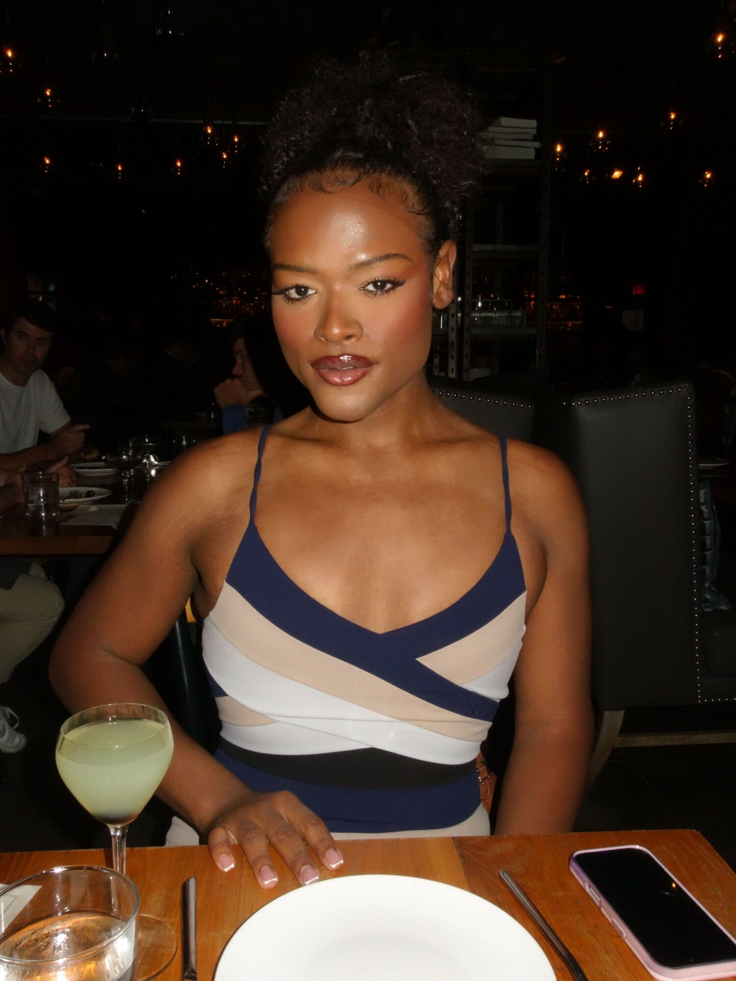 Woman with curly hair in a striped dress sitting at a table with drinks, nighttime setting.