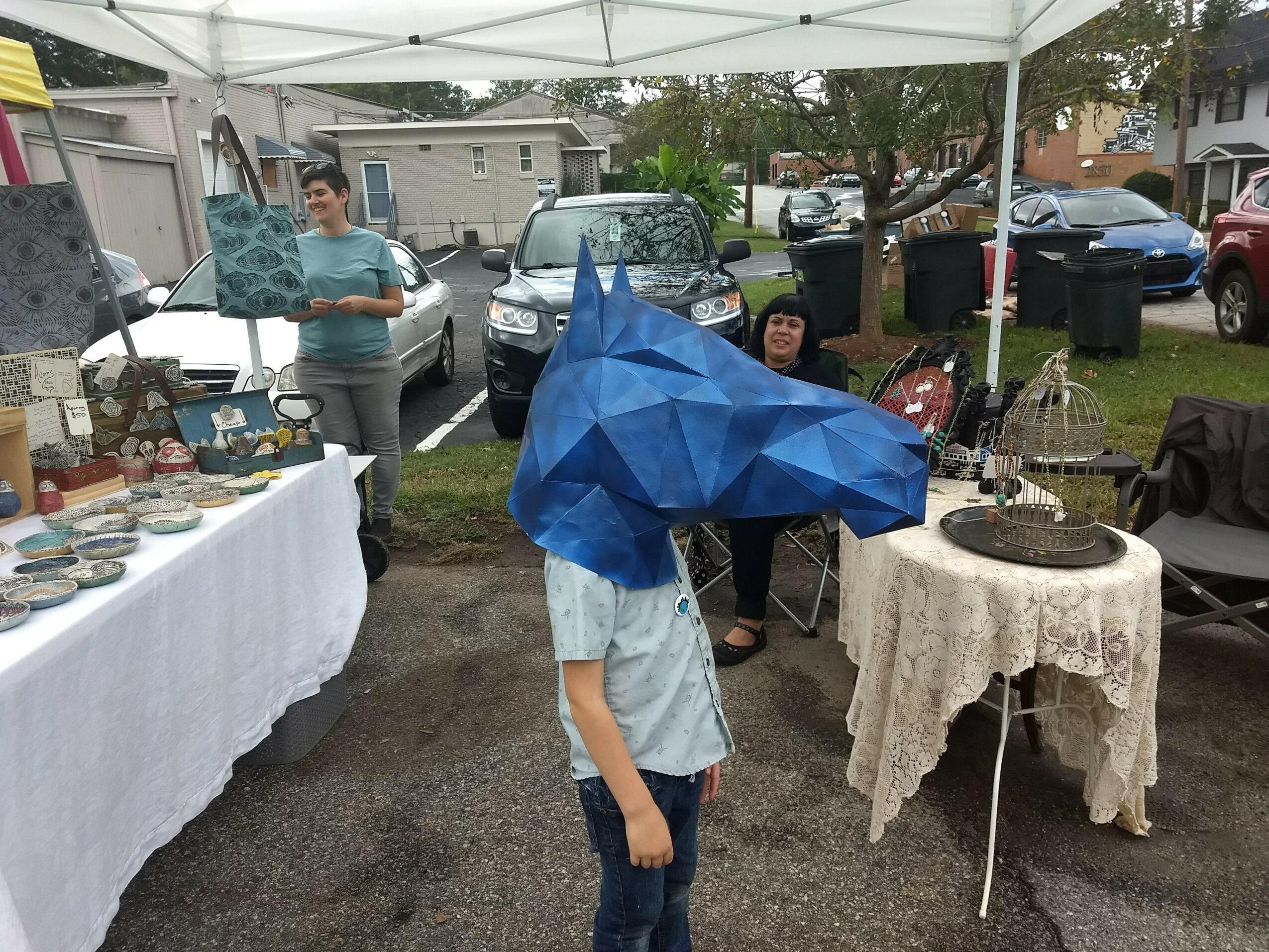 Person wearing a blue geometric animal mask standing outdoors near tables and cars under a canopy.