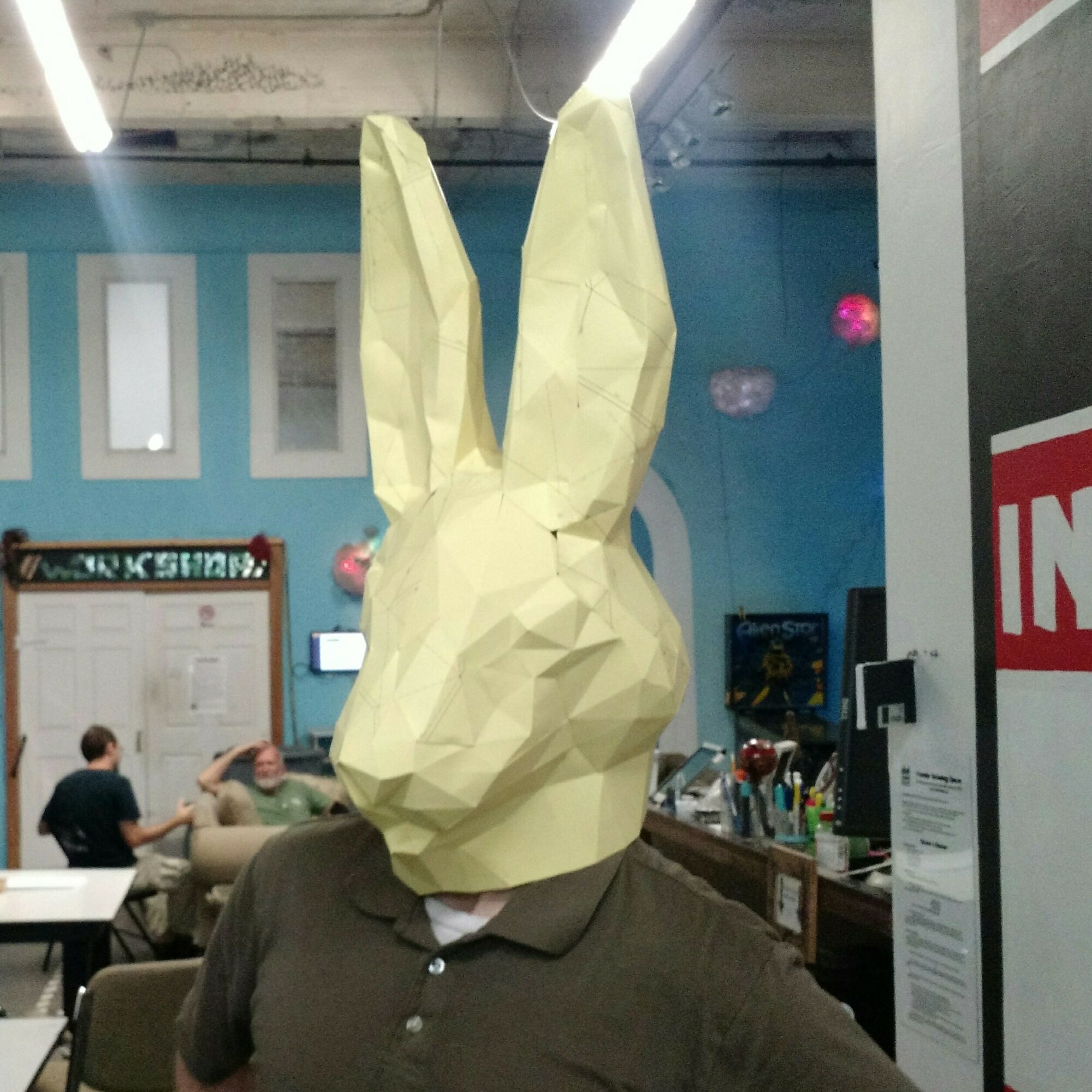 Person wearing a rabbit mask with tall ears in a room with people and blue walls.