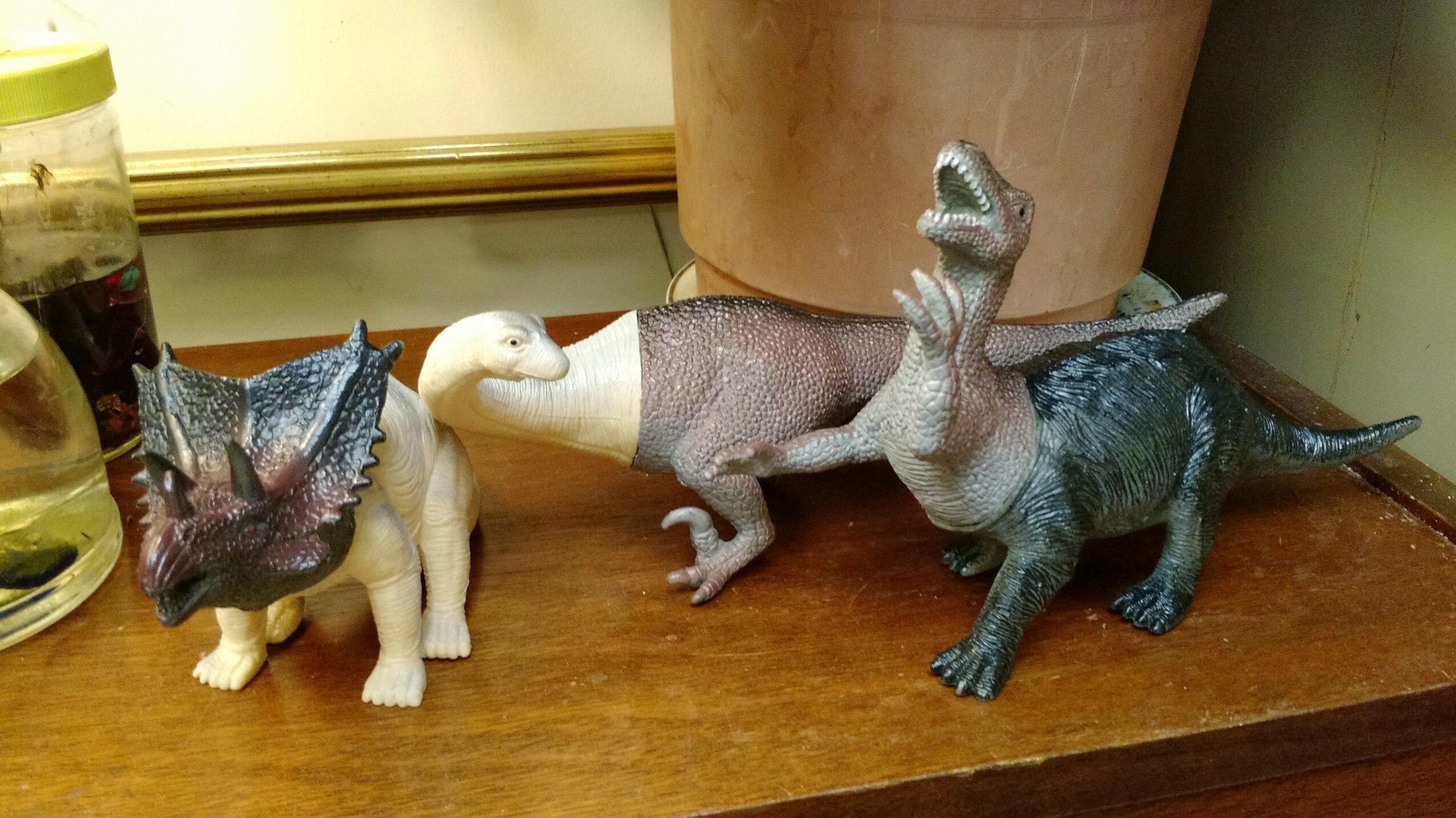 Three dinosaur figurines on a wooden surface, with a plant and a glass container in the background.
