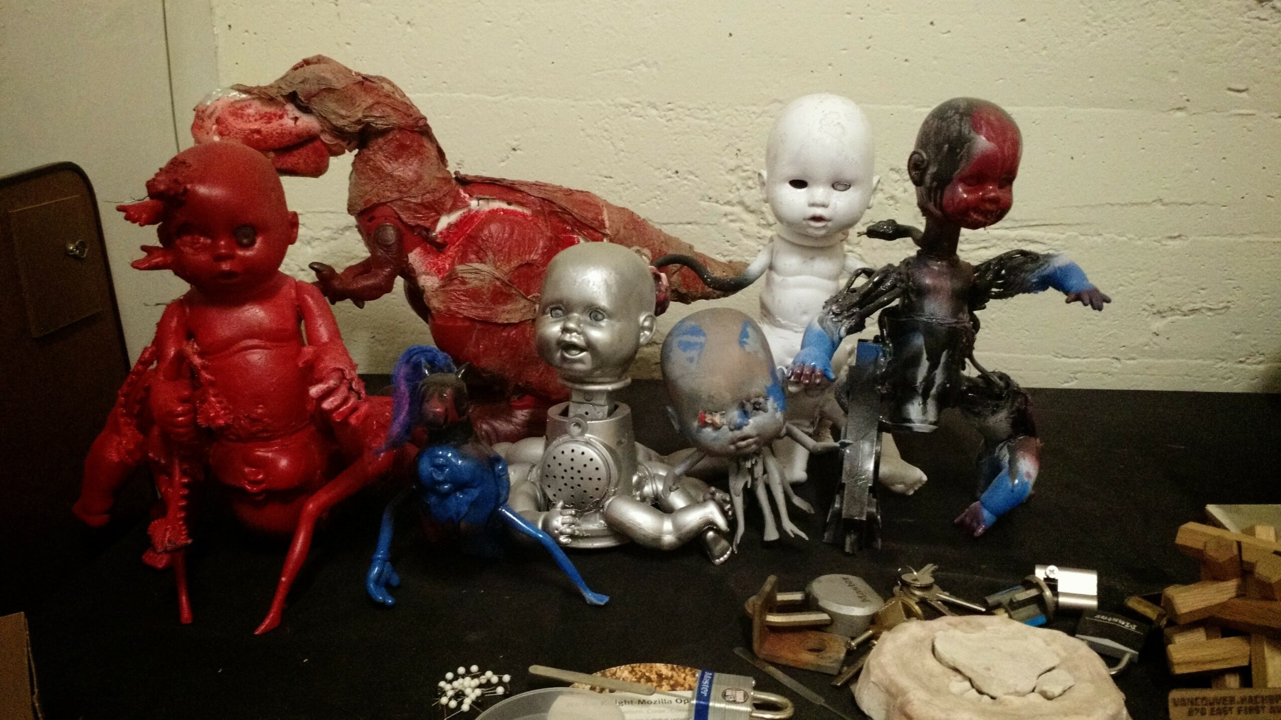 Collection of colorful dolls and figurines on a table with various objects nearby, against a plain wall.