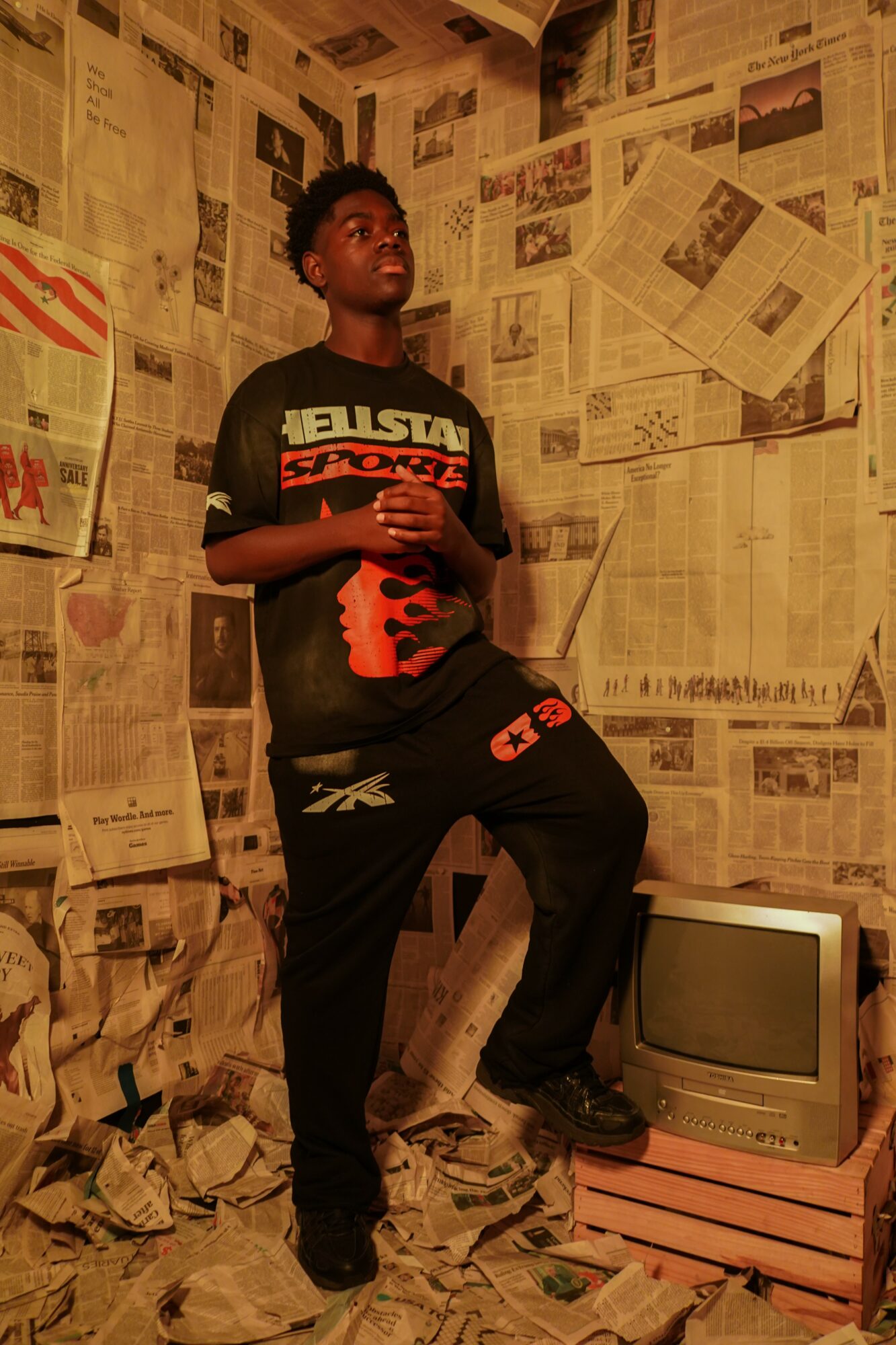 Young person standing in a room with walls covered in newspapers, next to an old television set, holding a phone.