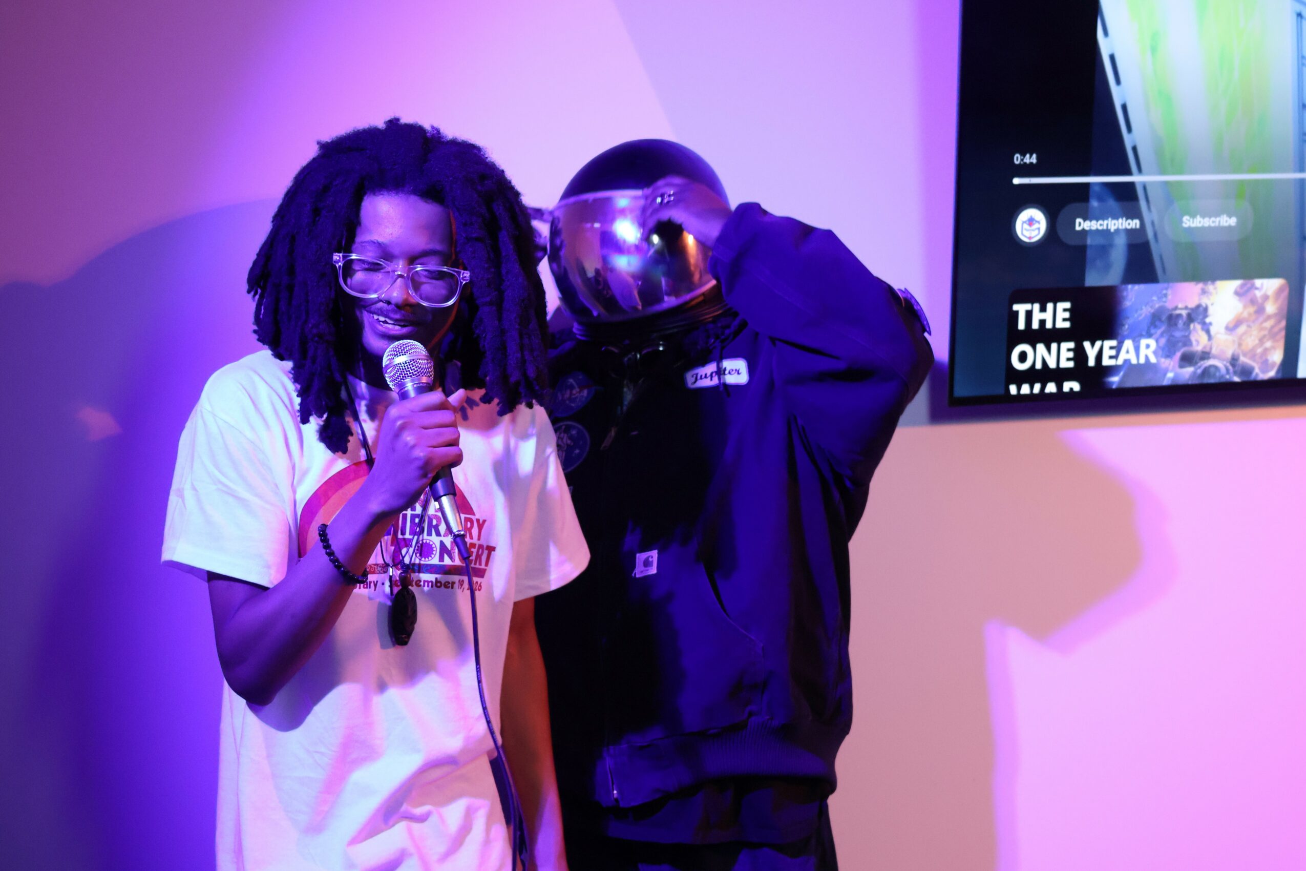 Two people on stage, one with glasses and dreadlocks holding a microphone, the other with a helmet, in front of a screen.