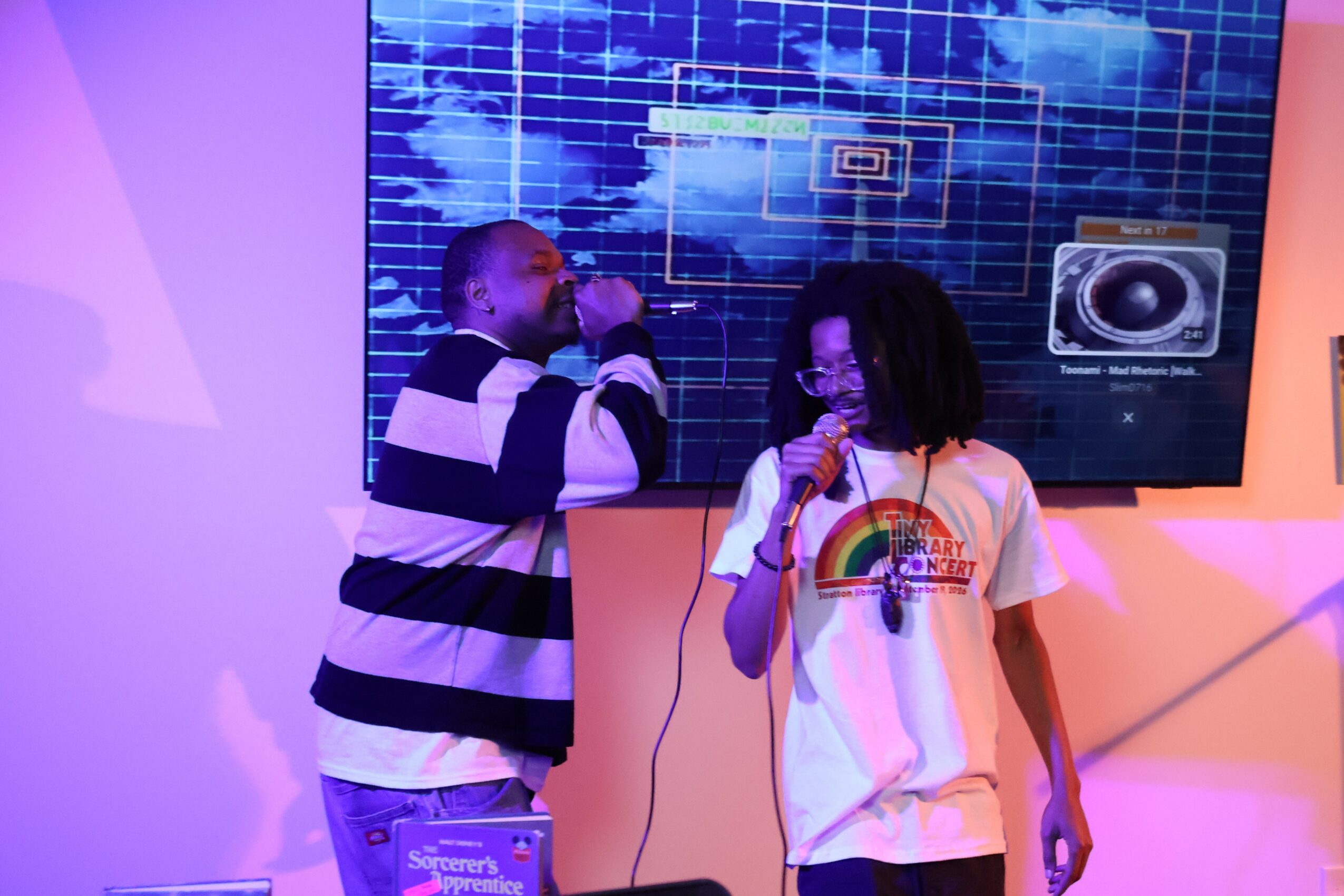 Two children singing into microphones on stage with a digital screen background, one wearing a striped shirt and the other a rainbow t-shirt.