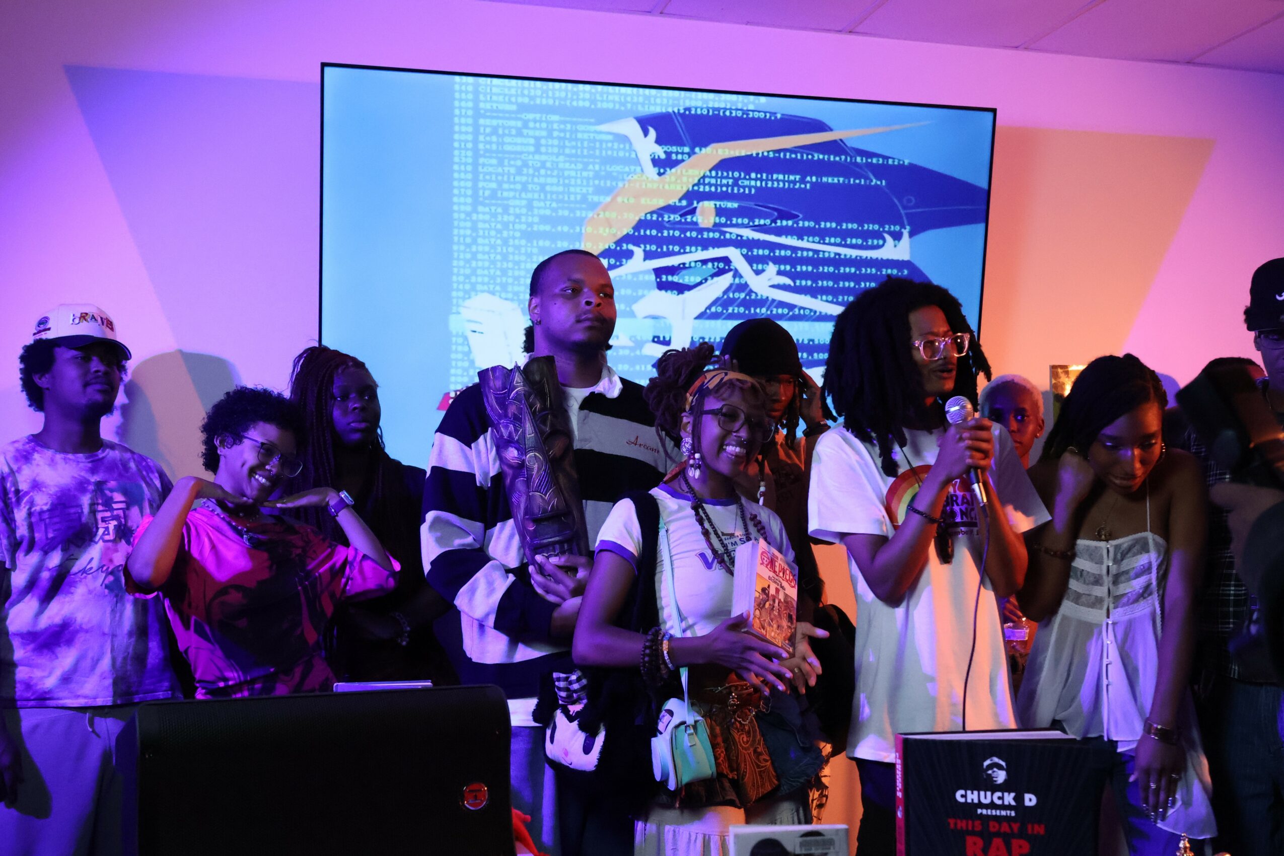 Group of young people standing on stage, some holding microphones, with a colorful background and a large screen behind them.