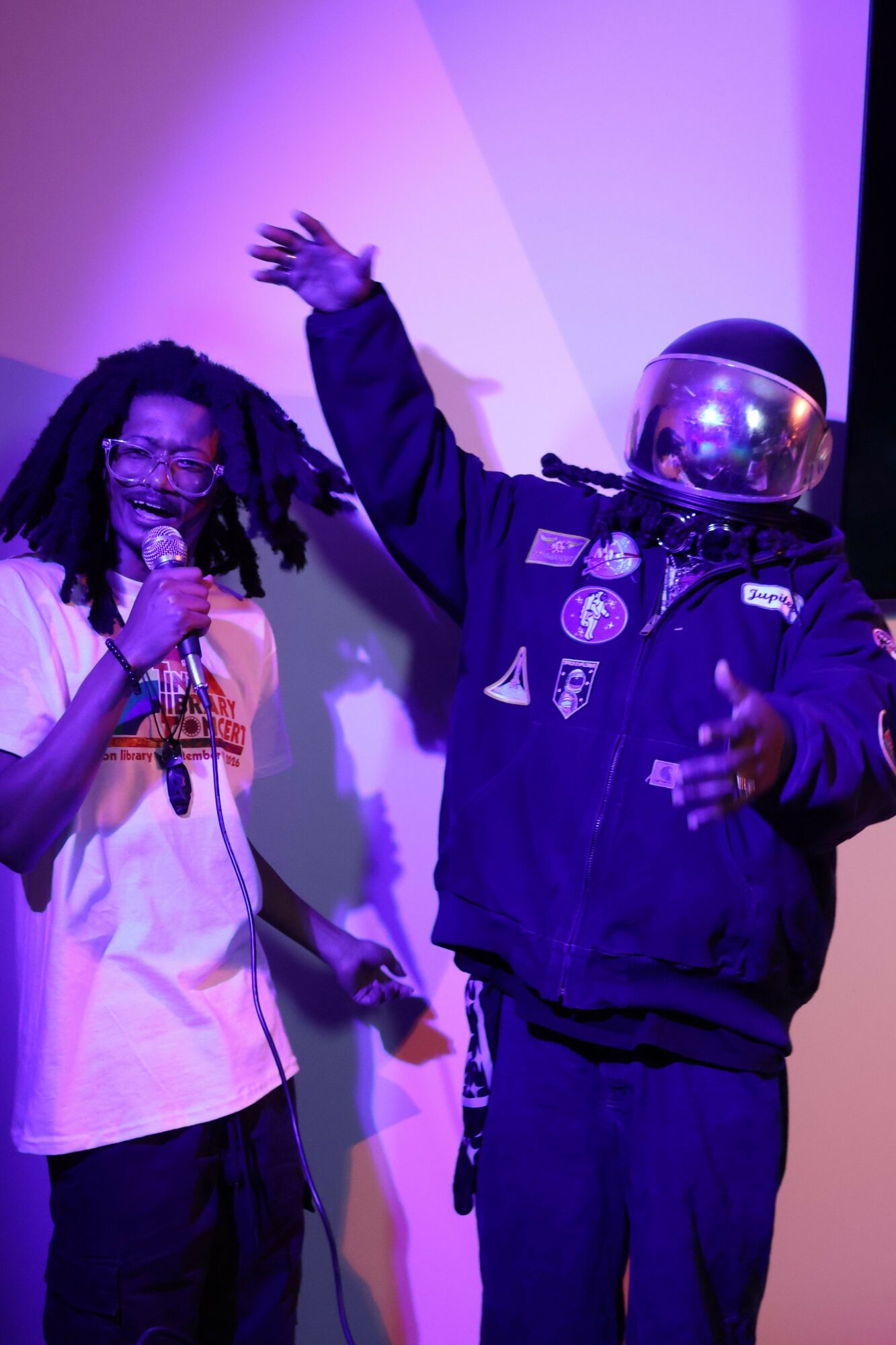 Person in police uniform with helmet, holding a microphone, raising hand, and another person in white coat with glasses, holding a microphone, in a room with purple lighting.