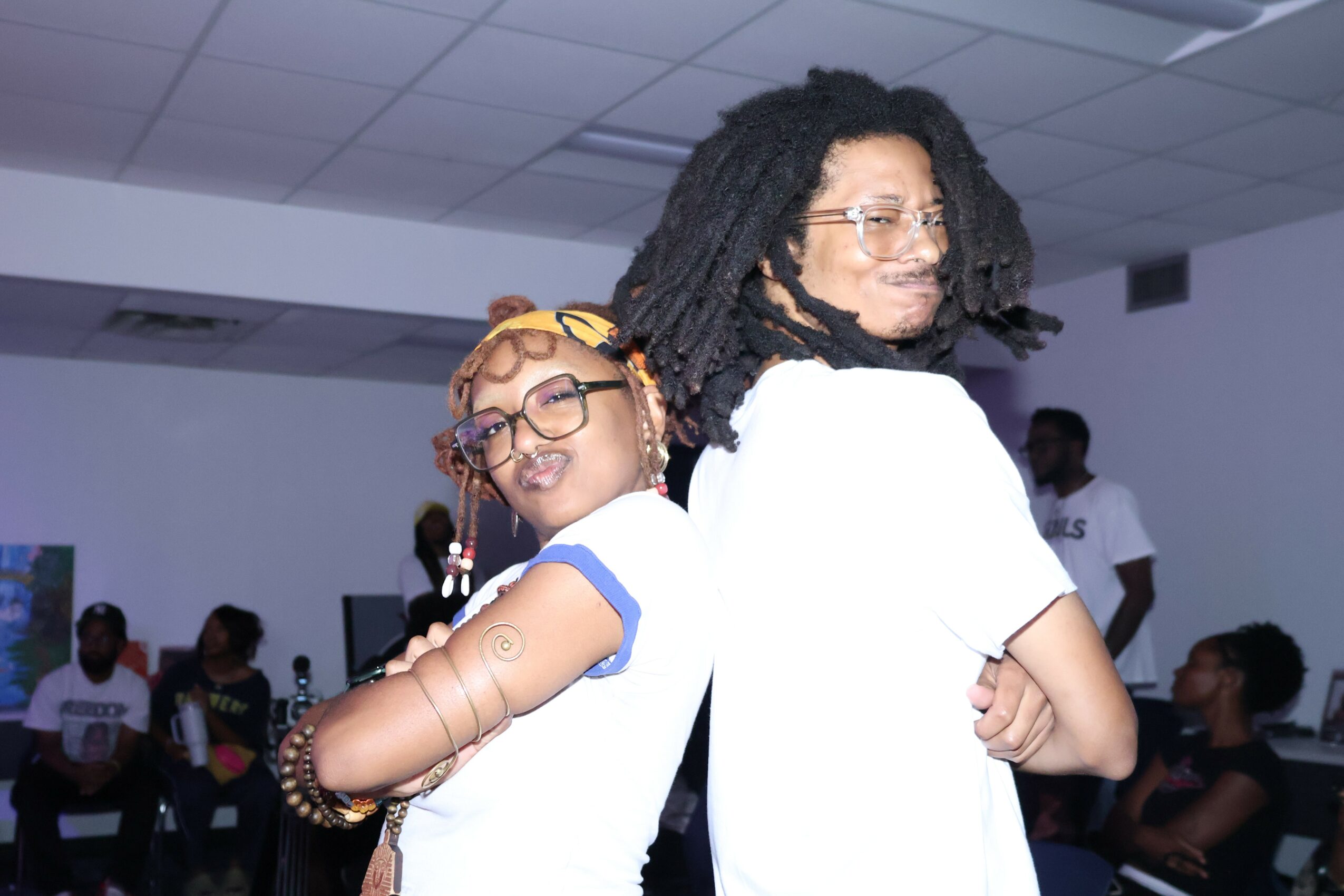 Two people standing back-to-back, smiling, in a room with other people in the background.