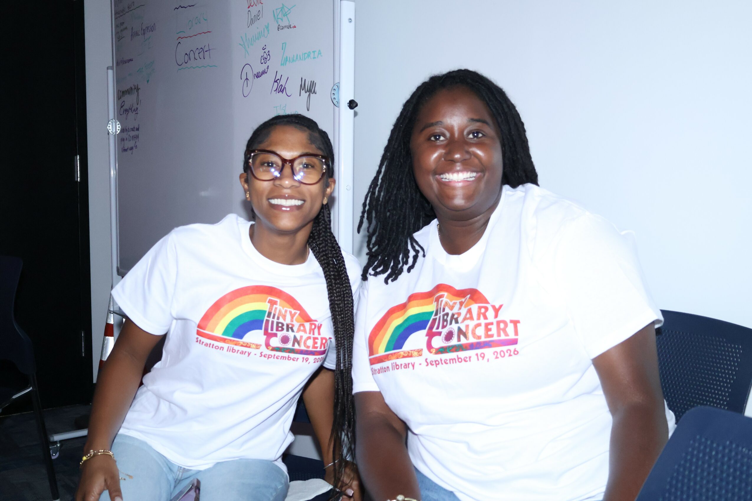 Two women smiling, wearing matching white T-shirts with rainbow and text, sitting indoors against a white wall.