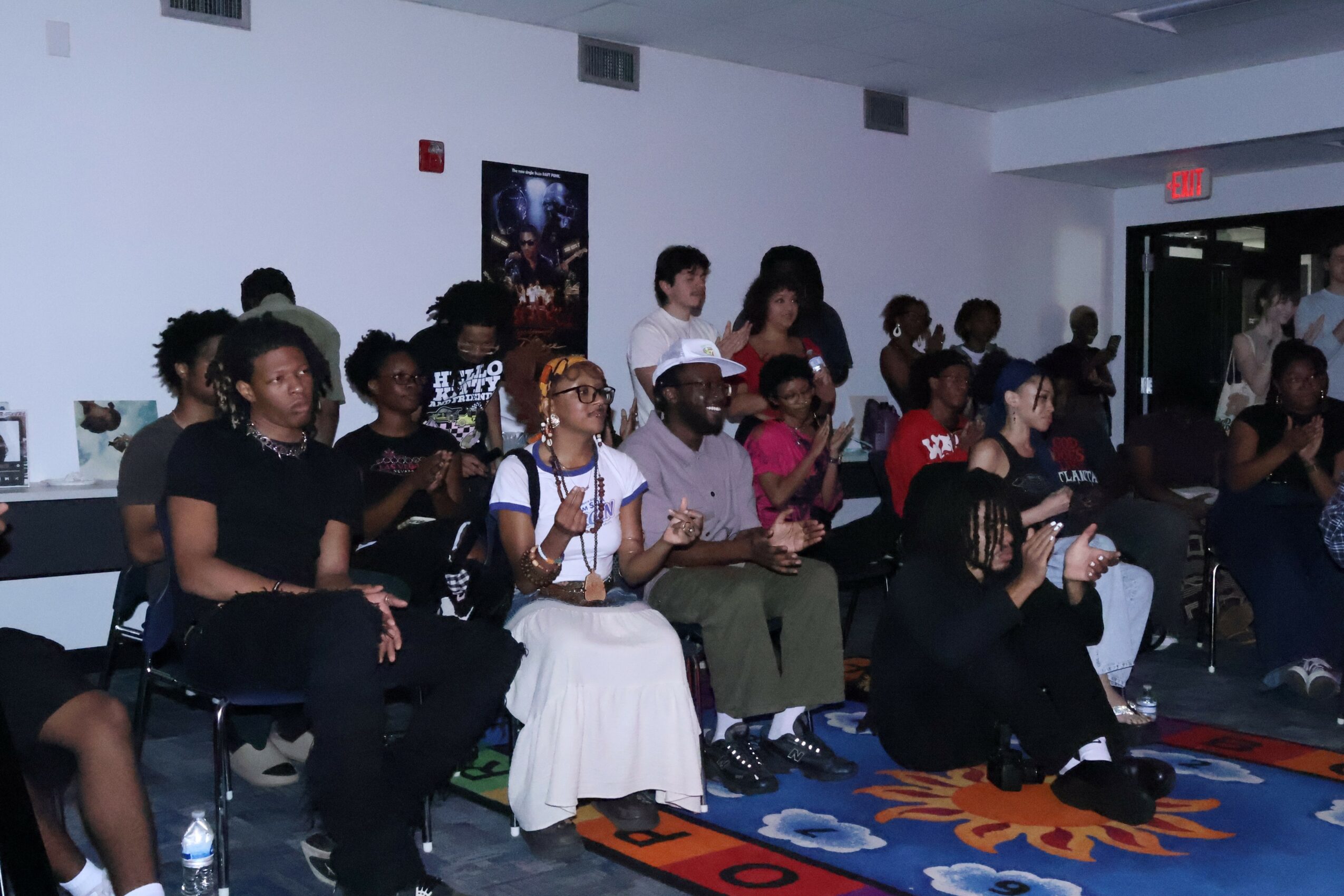 Group of people seated indoors, some looking forward, others engaged in conversation, with a colorful rug on the floor.
