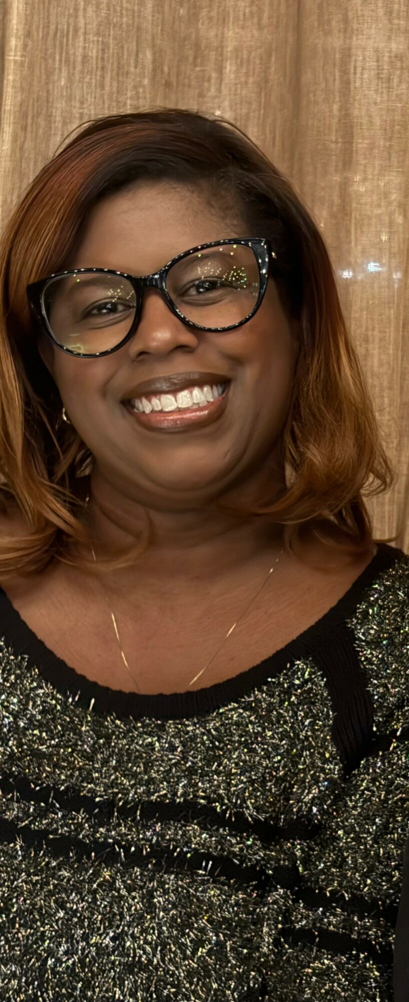Smiling woman with glasses and shoulder-length hair, wearing a sparkly top, in front of a wooden background.