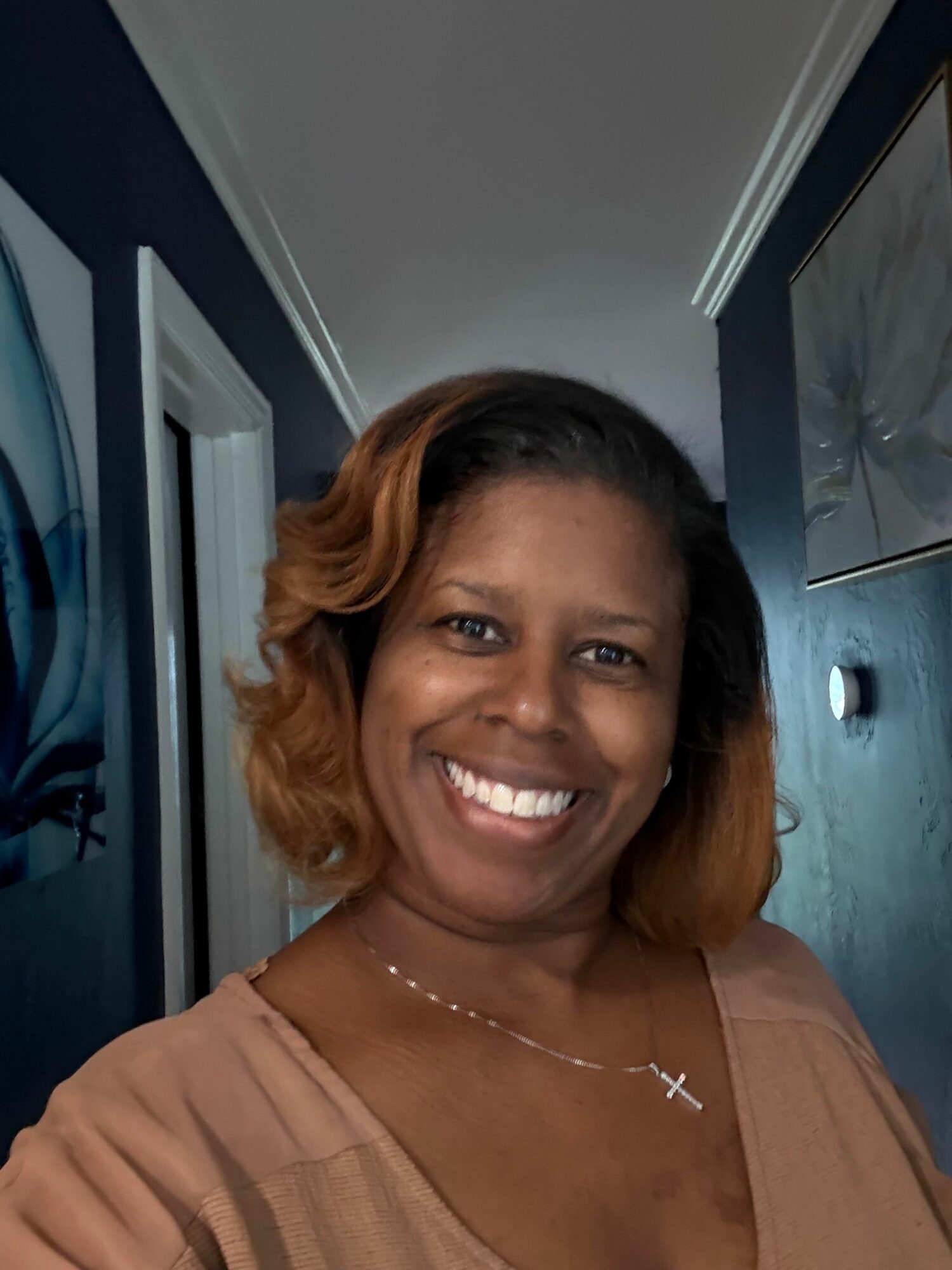 Smiling woman with shoulder-length hair, wearing a necklace and a beige top, in a room with a door and a painting behind her.