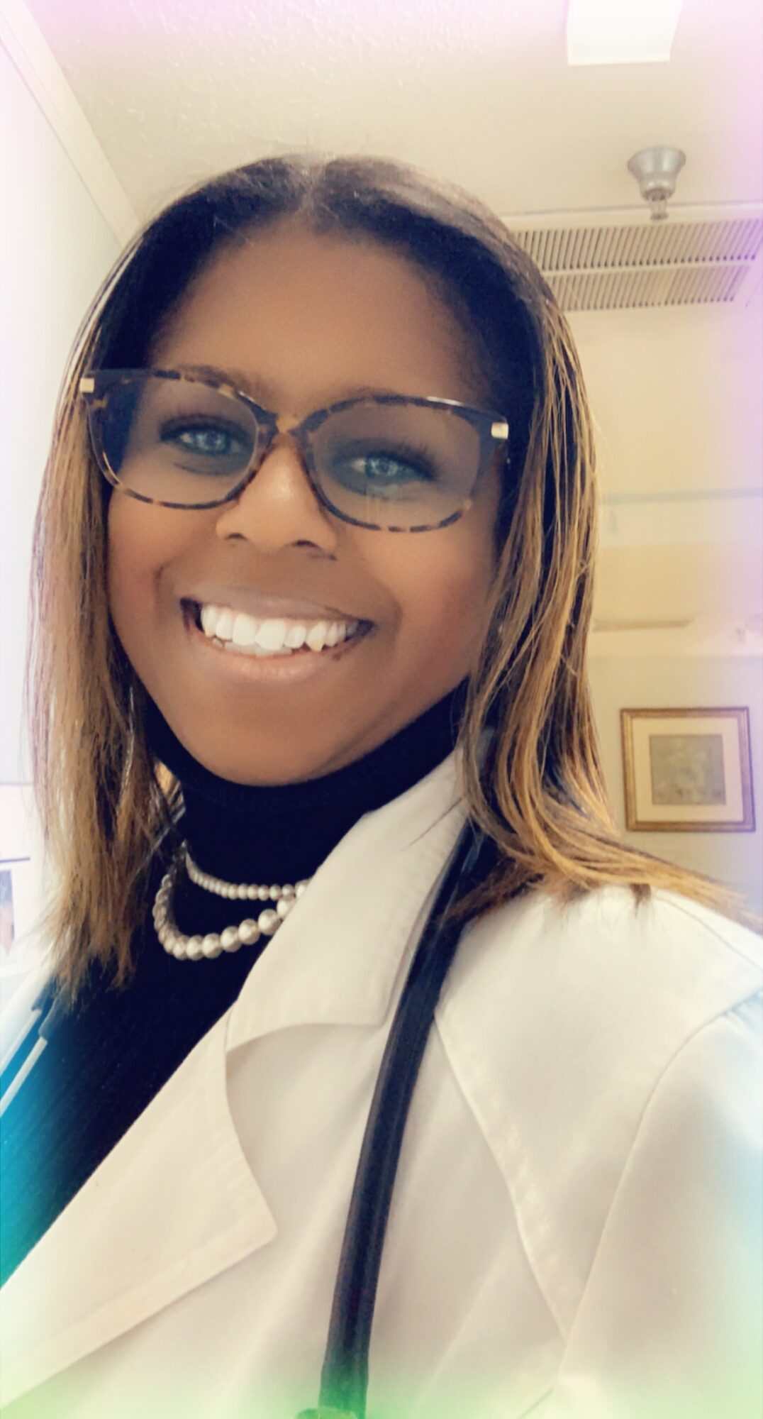 Smiling woman wearing glasses, white coat, and pearl necklace, with a stethoscope, in a professional setting.