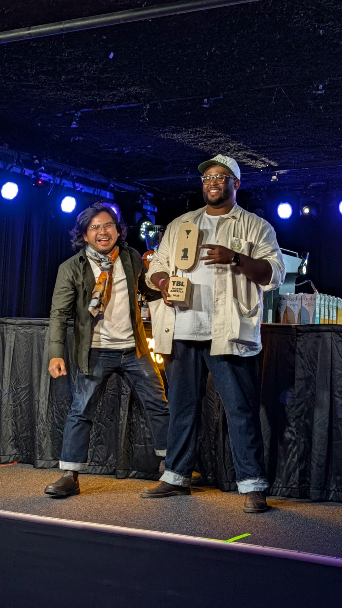 Two people standing on stage under blue lights, one holding a trophy and a certificate, smiling.
