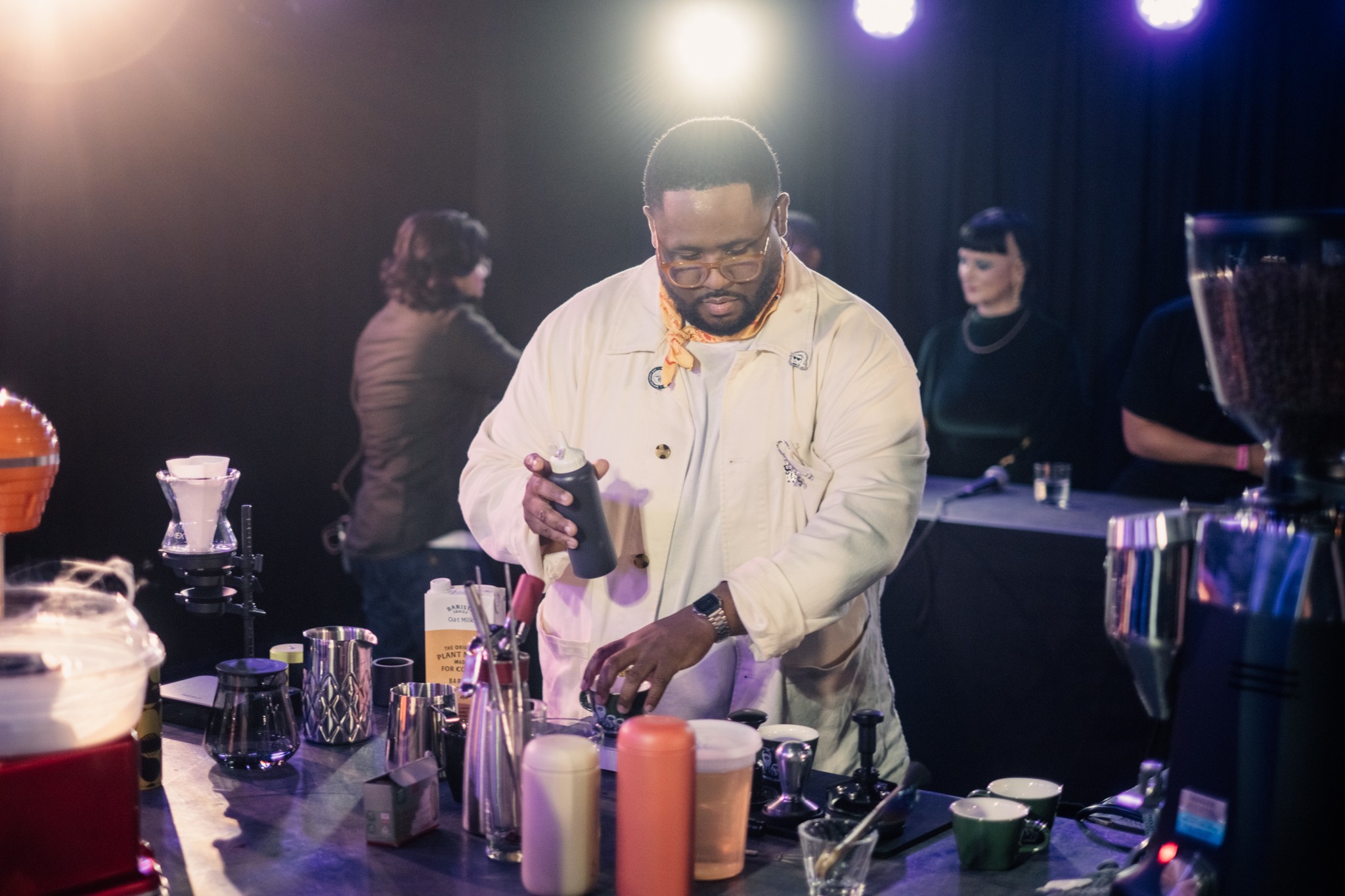 Man preparing drinks at a bar with various bottles and bar tools, with people in the background, dim lighting, and spotlights.
