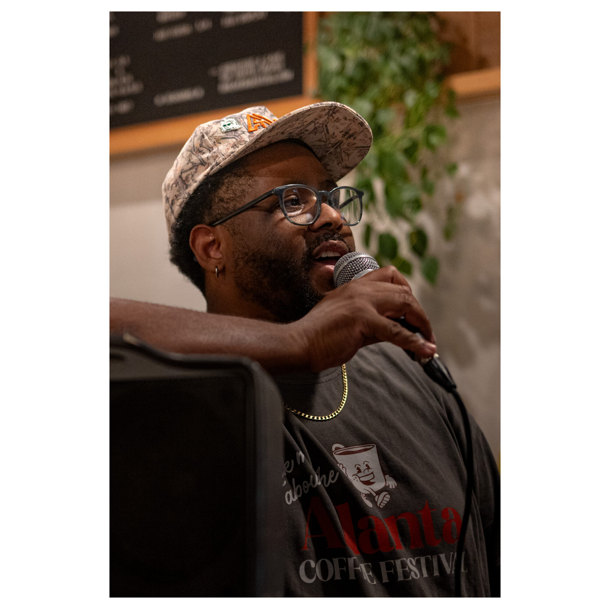 Person speaking into a microphone, wearing glasses and a cap, with a blurred background and a plant. 20 words.