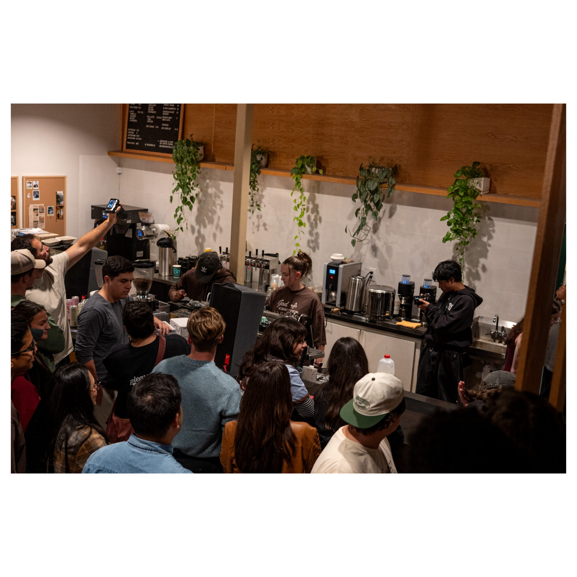 Crowd gathered in a coffee shop, baristas preparing drinks behind the counter, green plants hanging on the wall.