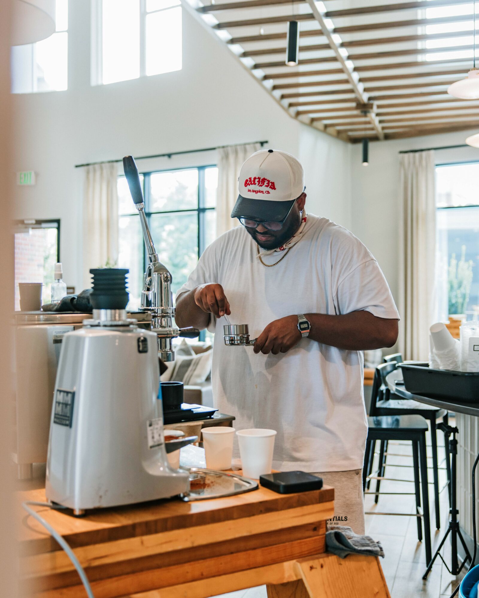 Person wearing a white t-shirt, cap, and glasses preparing coffee in a bright, modern cafe with large windows and wooden ceiling.