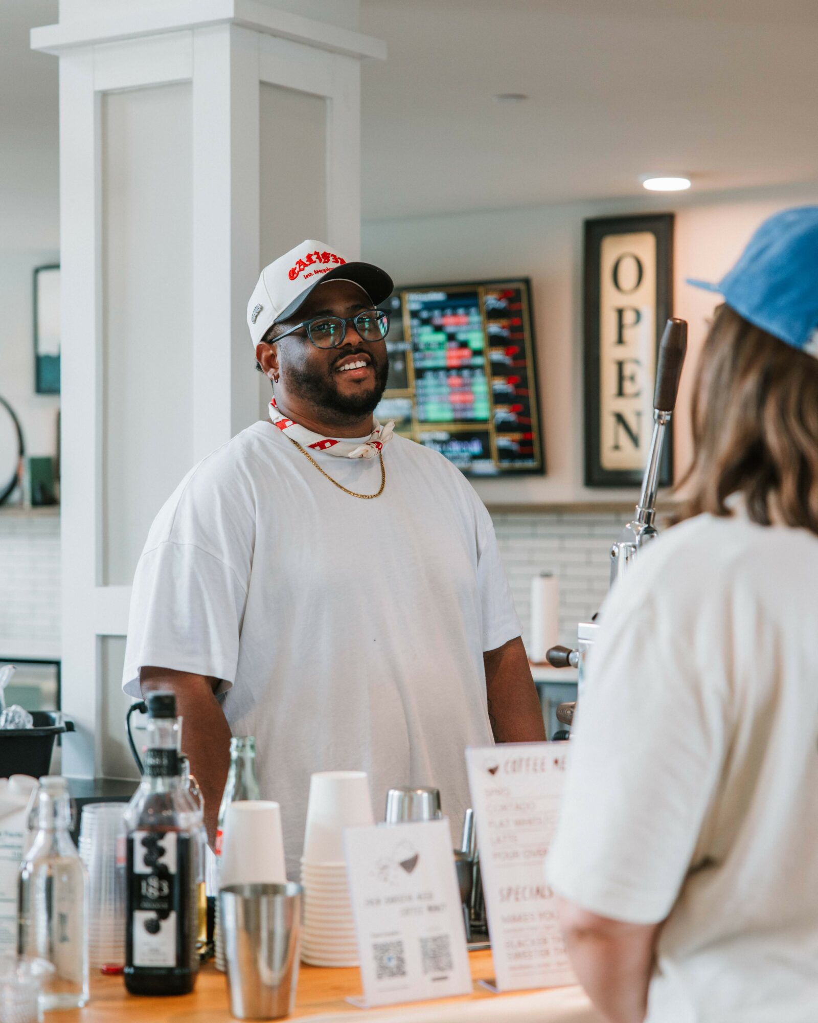 Smiling man with glasses and a cap behind a counter, with a person in a blue cap in front, in a cafe or shop setting.