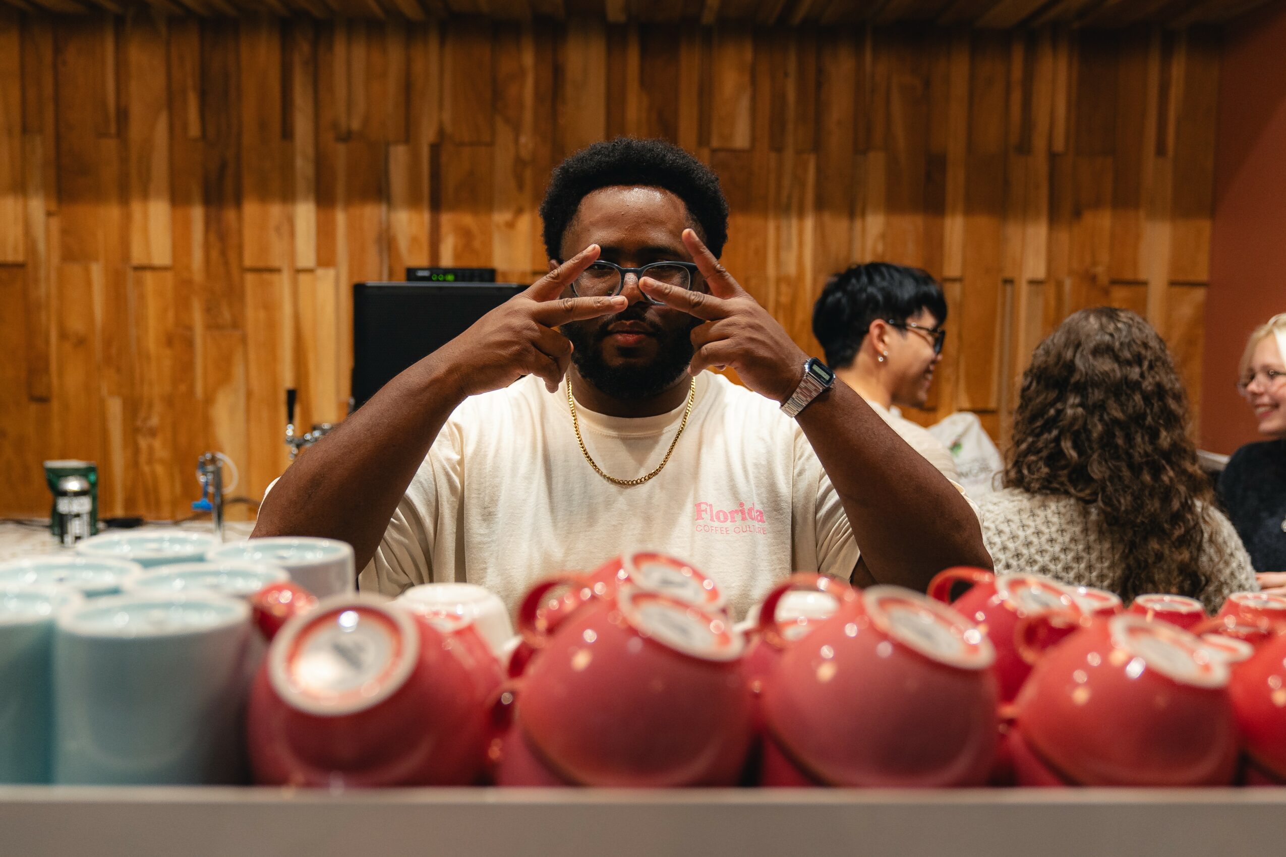 Person with glasses and a beard making a gesture with hands near face, surrounded by colorful cups, in a room with wooden walls.