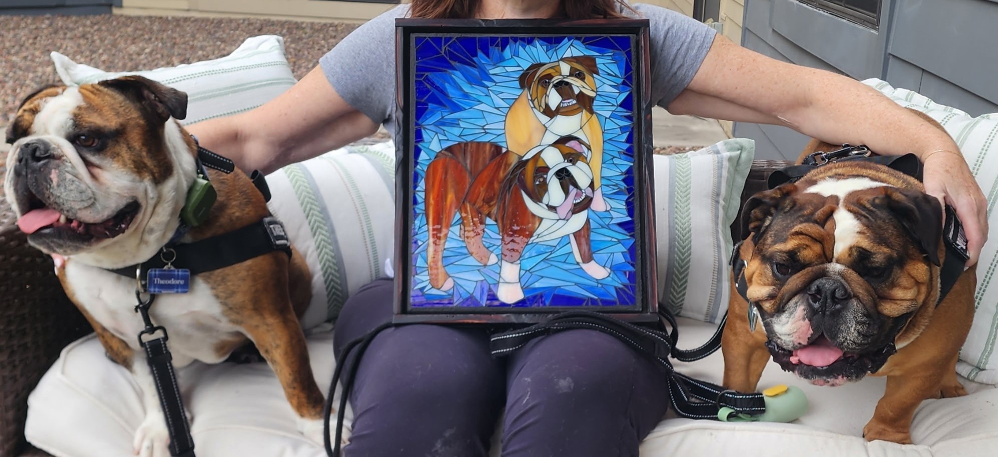 Person sitting on a couch with two bulldogs, holding a framed picture of a dog, in a living room setting.