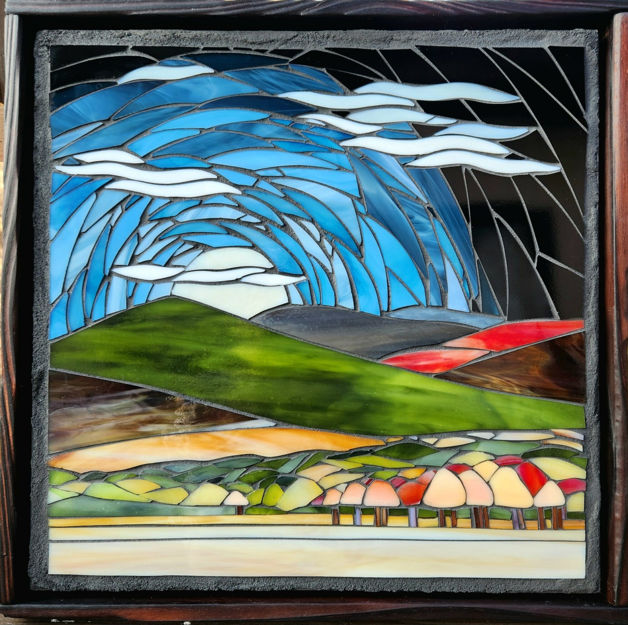 Colorful stained glass landscape with sky, clouds, green hills, and a row of houses, in a square frame.