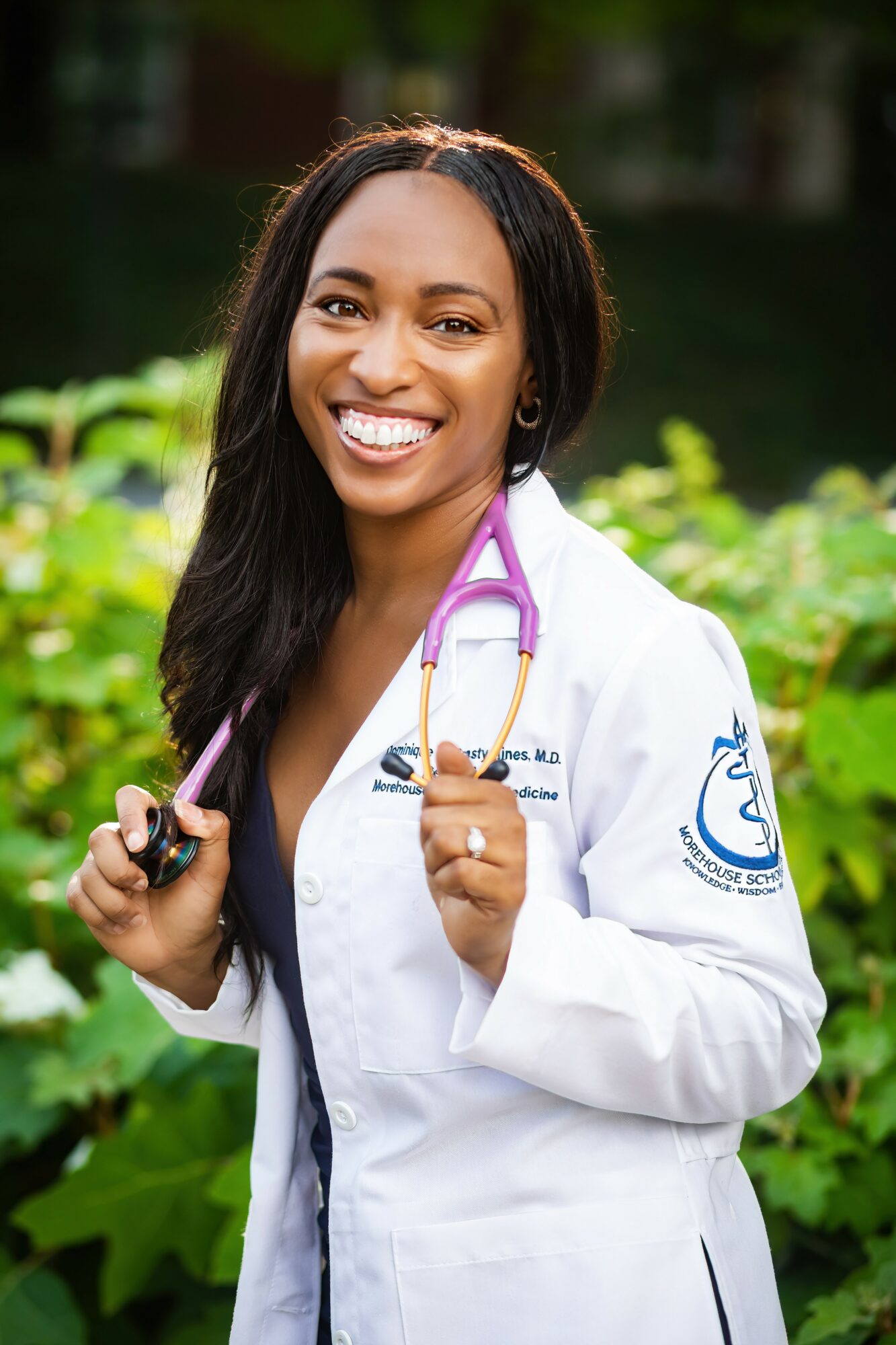 Smiling female doctor outdoors holding a stethoscope, wearing a white coat with a medical emblem, green background.