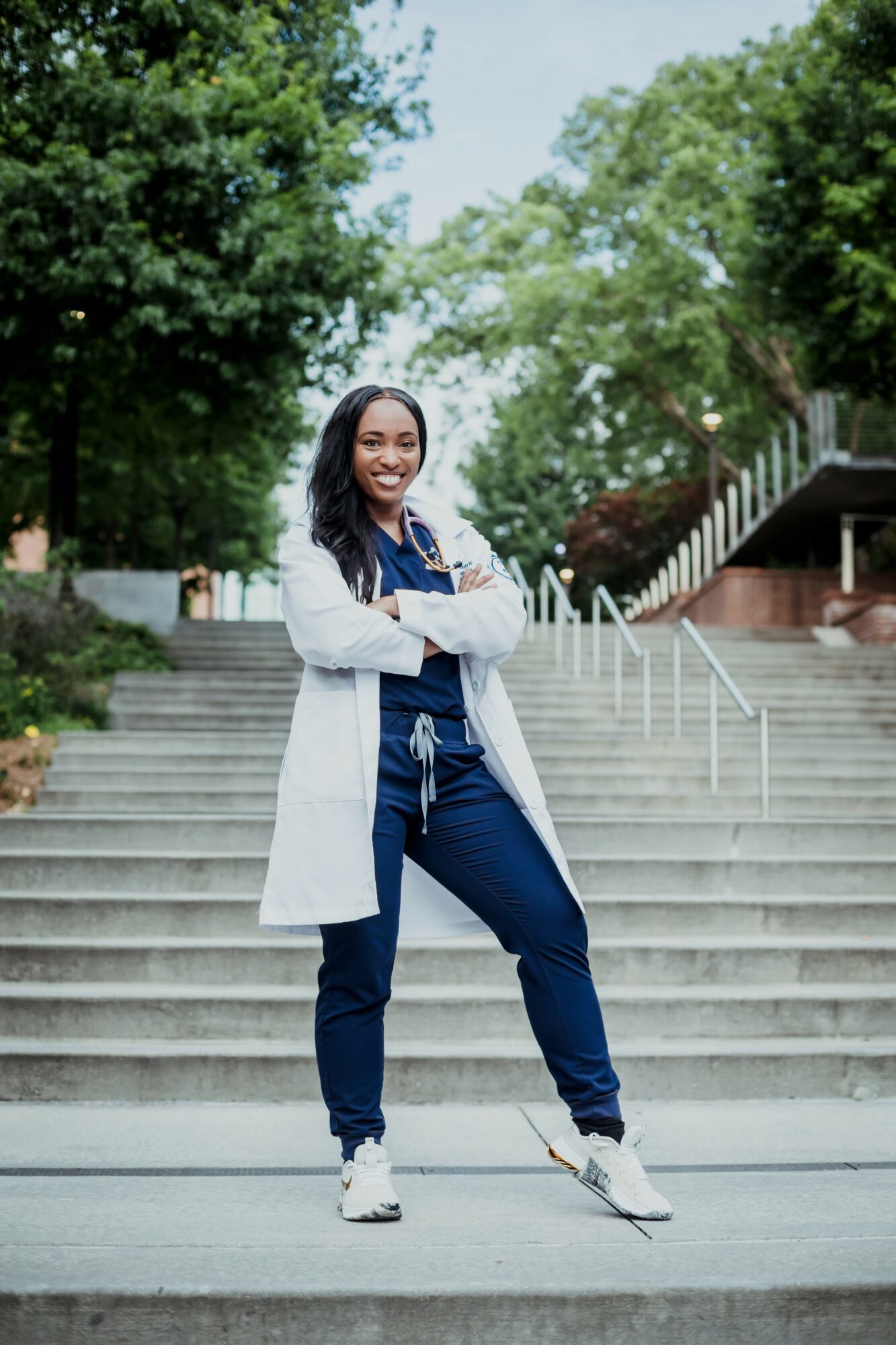 Woman standing on outdoor stairs with trees in background, smiling, arms crossed, wearing a white coat and jeans.